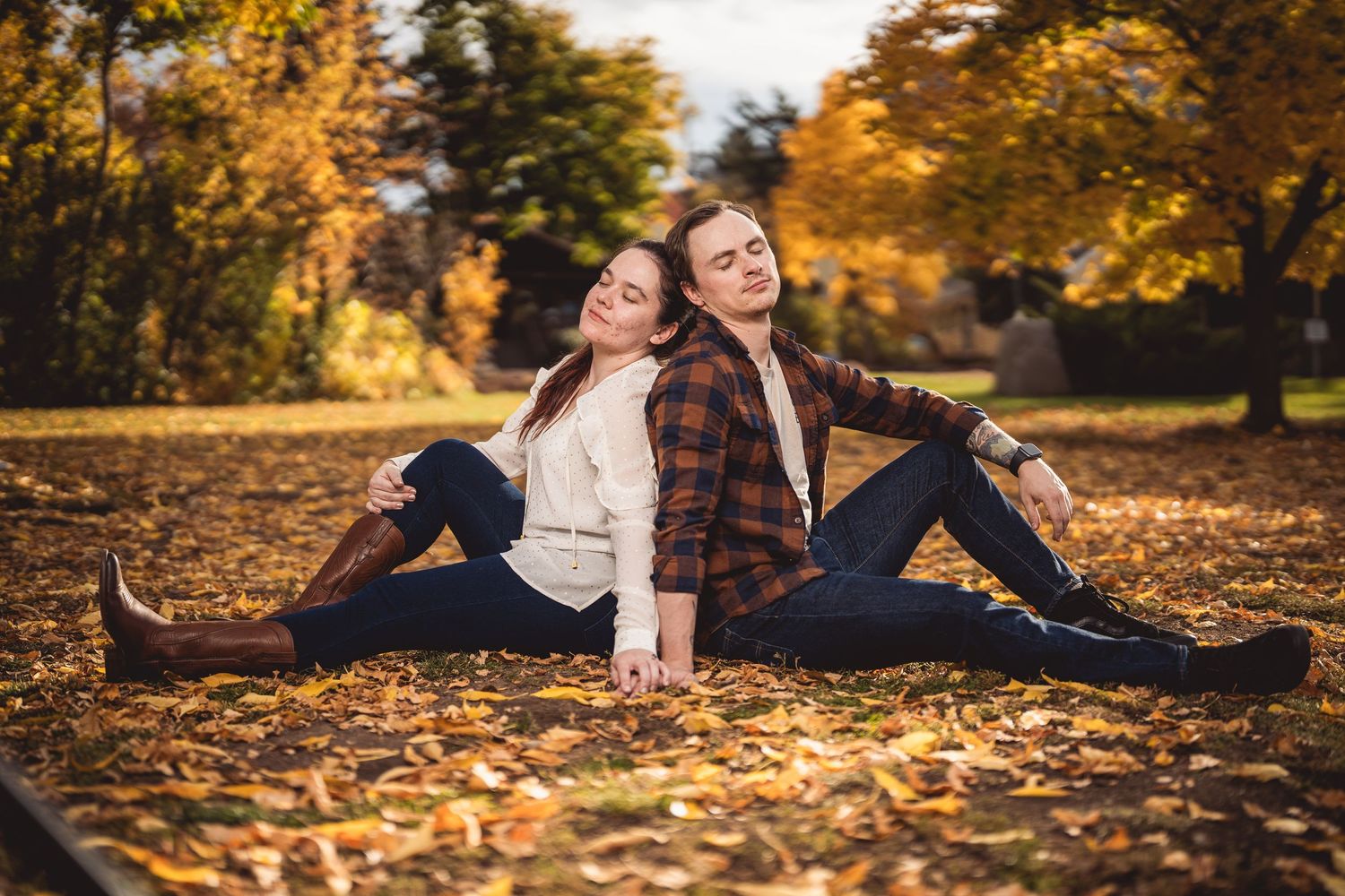 Couple sitting back-to-back on autumn leaves in a park, surrounded by golden foliage in a romantic fall setting.