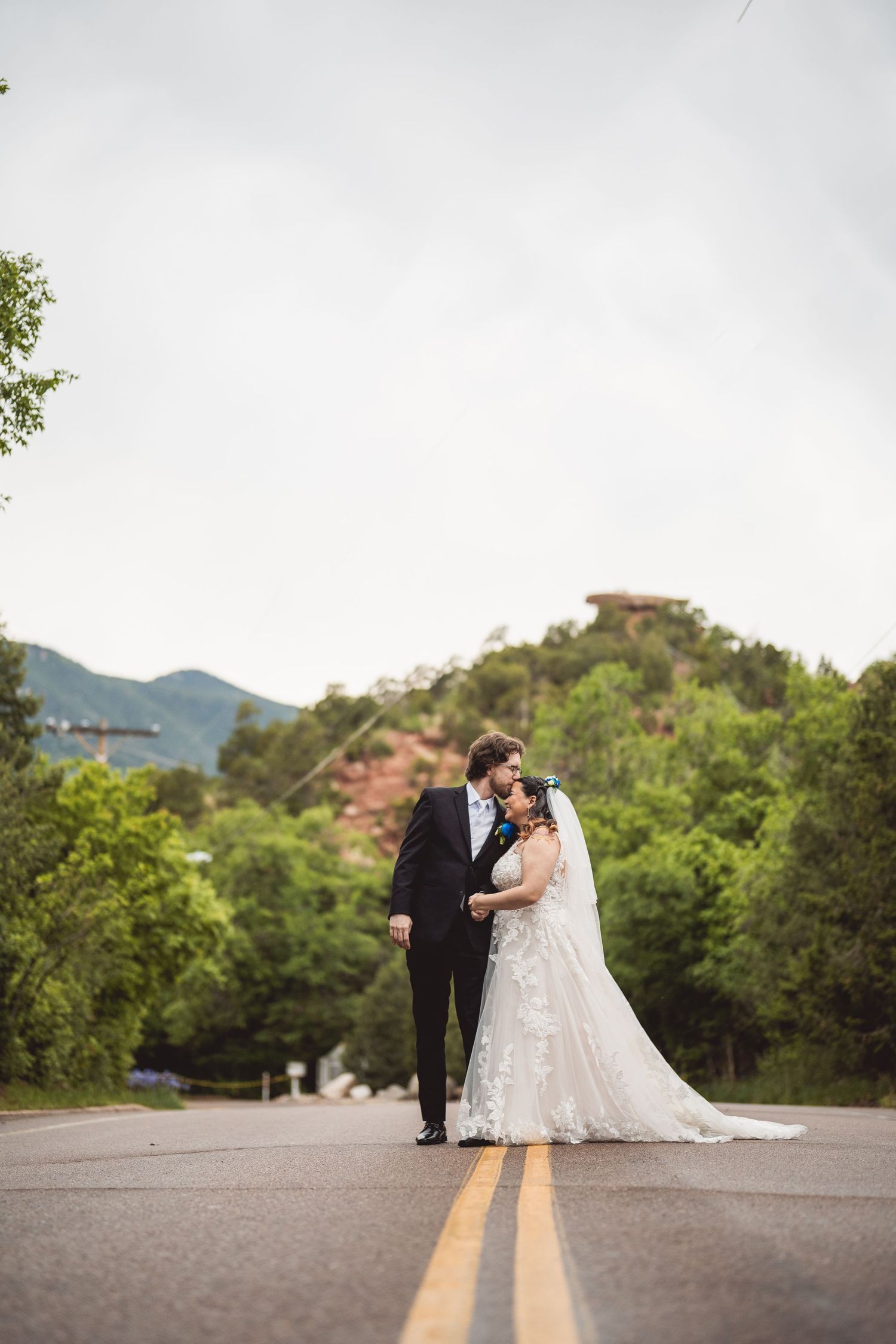 Bride and groom share a romantic moment on a mountain road with lush green trees and red rock formations behind them.