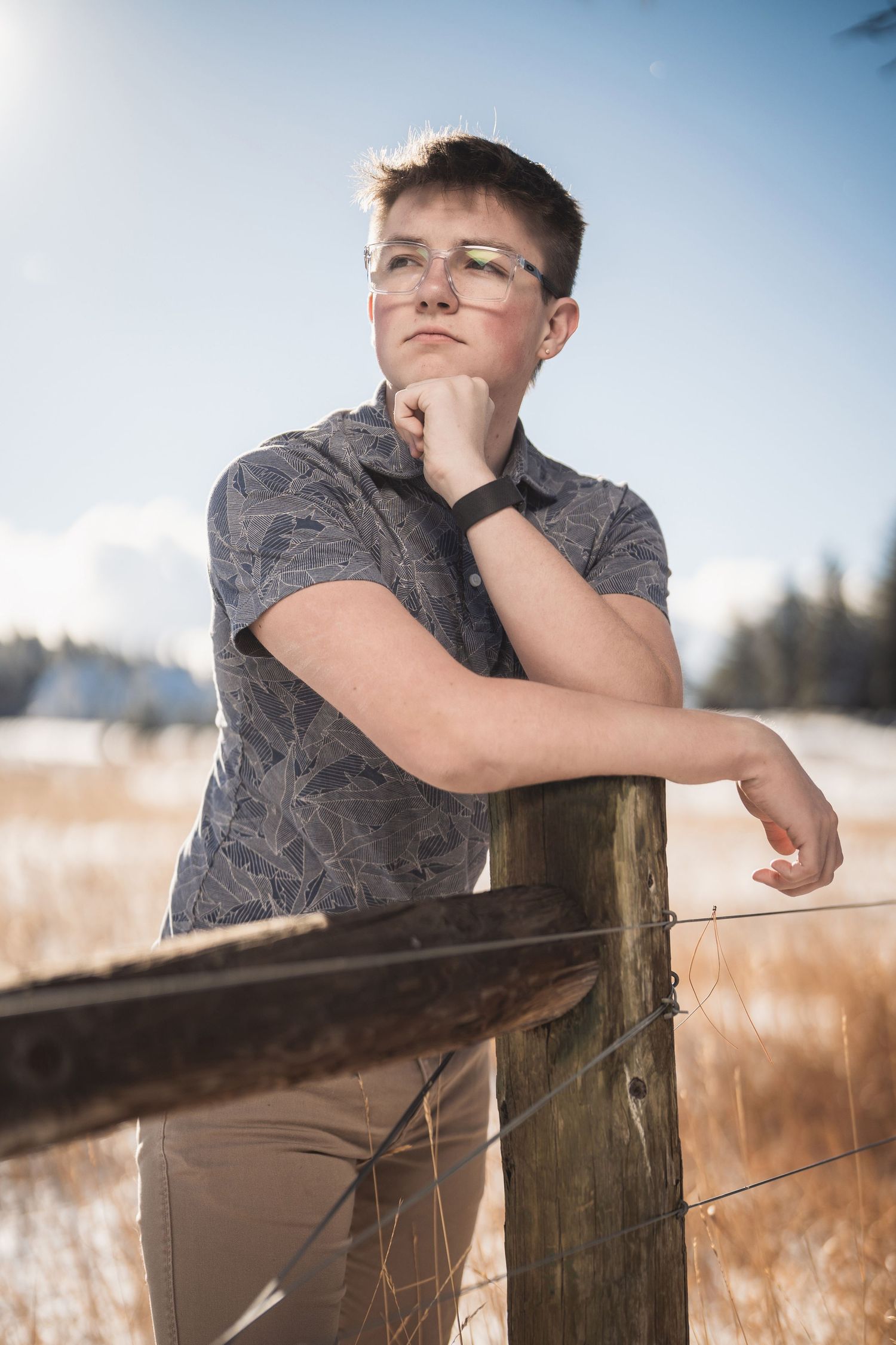 Young person with glasses leaning on a wooden fence post in a snowy field, wearing a gray patterned shirt.