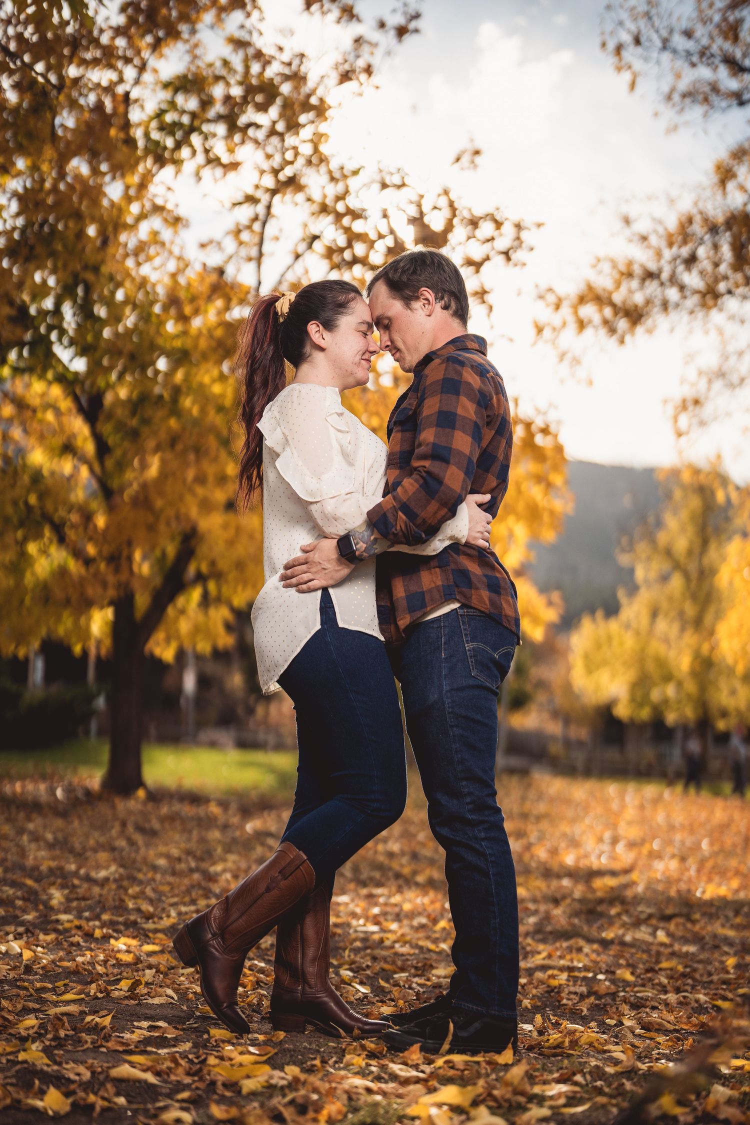 Couple in fall attire embracing among golden autumn leaves on ground.