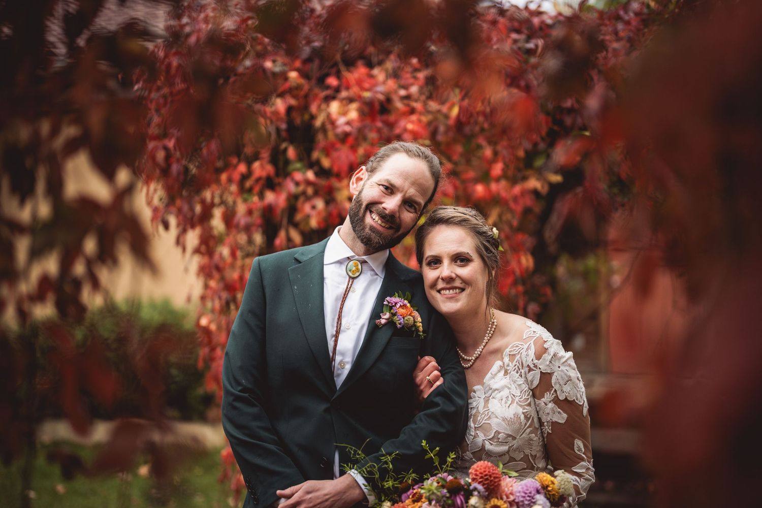 Newlywed couple sharing a joyful moment amidst vibrant red autumn foliage with beautiful floral bouquet.