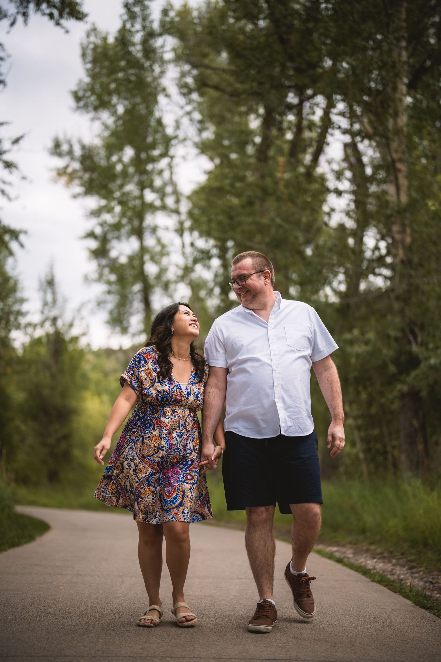 A couple takes a leisurely stroll together on a paved walking path surrounded by trees.