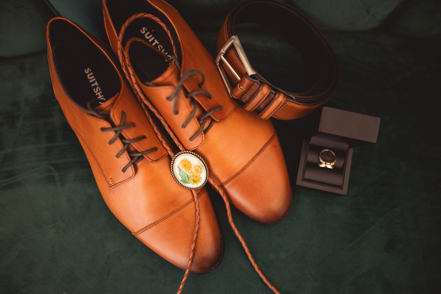 Brown leather dress shoes and wedding rings arranged on a dark surface.