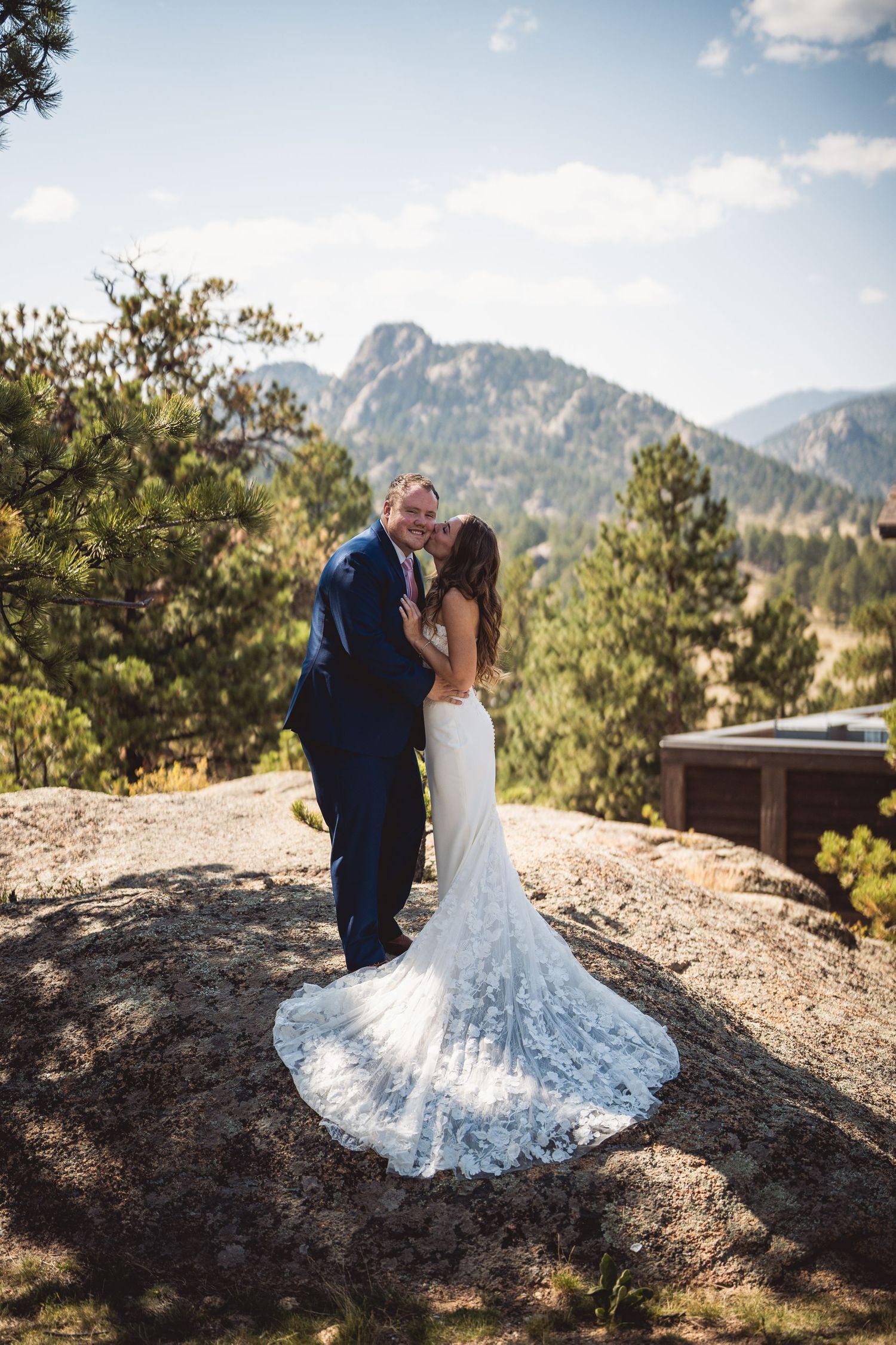 Bride and groom embrace on rocky mountain overlook, lace wedding gown train spread, pine trees and peaks behind.