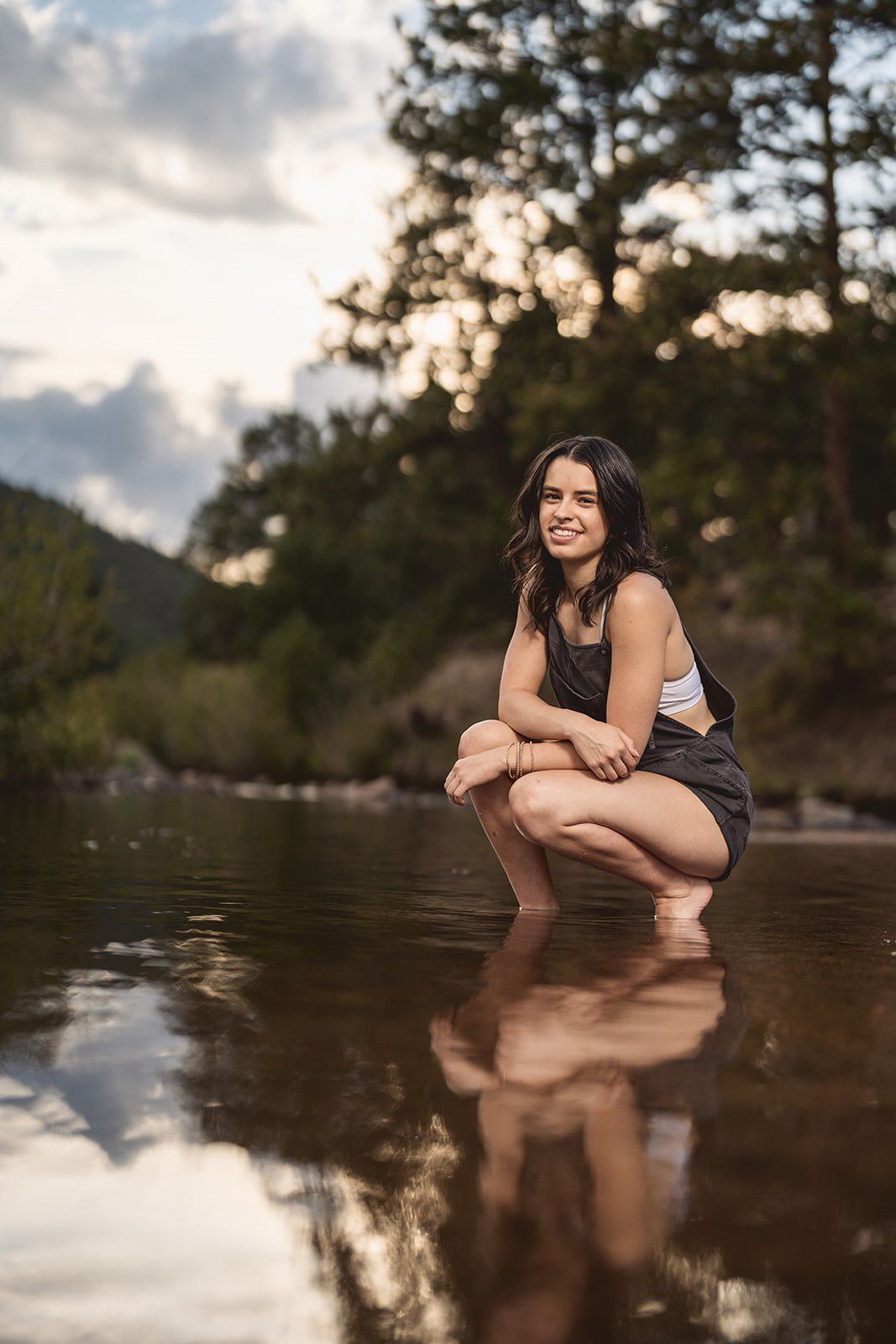 Young woman crouching in shallow river water smiling, surrounded by trees and cloudy sky reflected in water.