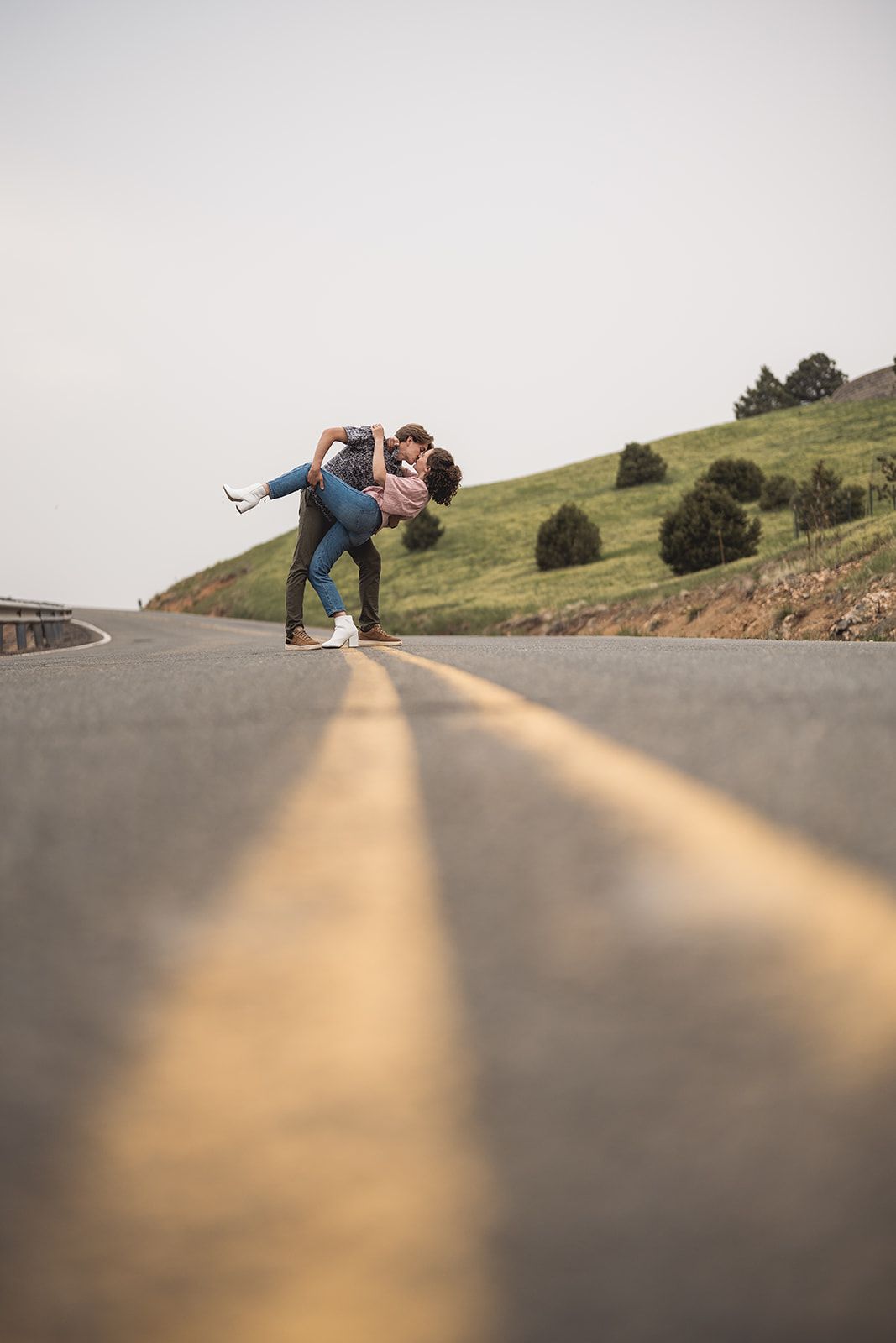 Couple sharing a romantic kiss on an empty road with yellow center lines and green hills in the background.