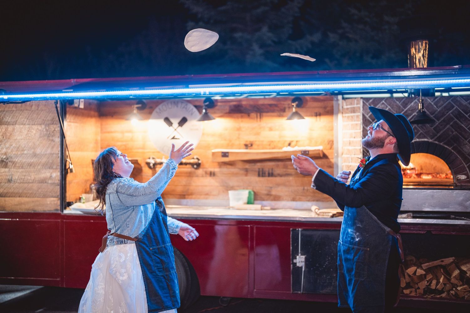 Newlyweds having fun tossing pizza dough at illuminated food truck station.