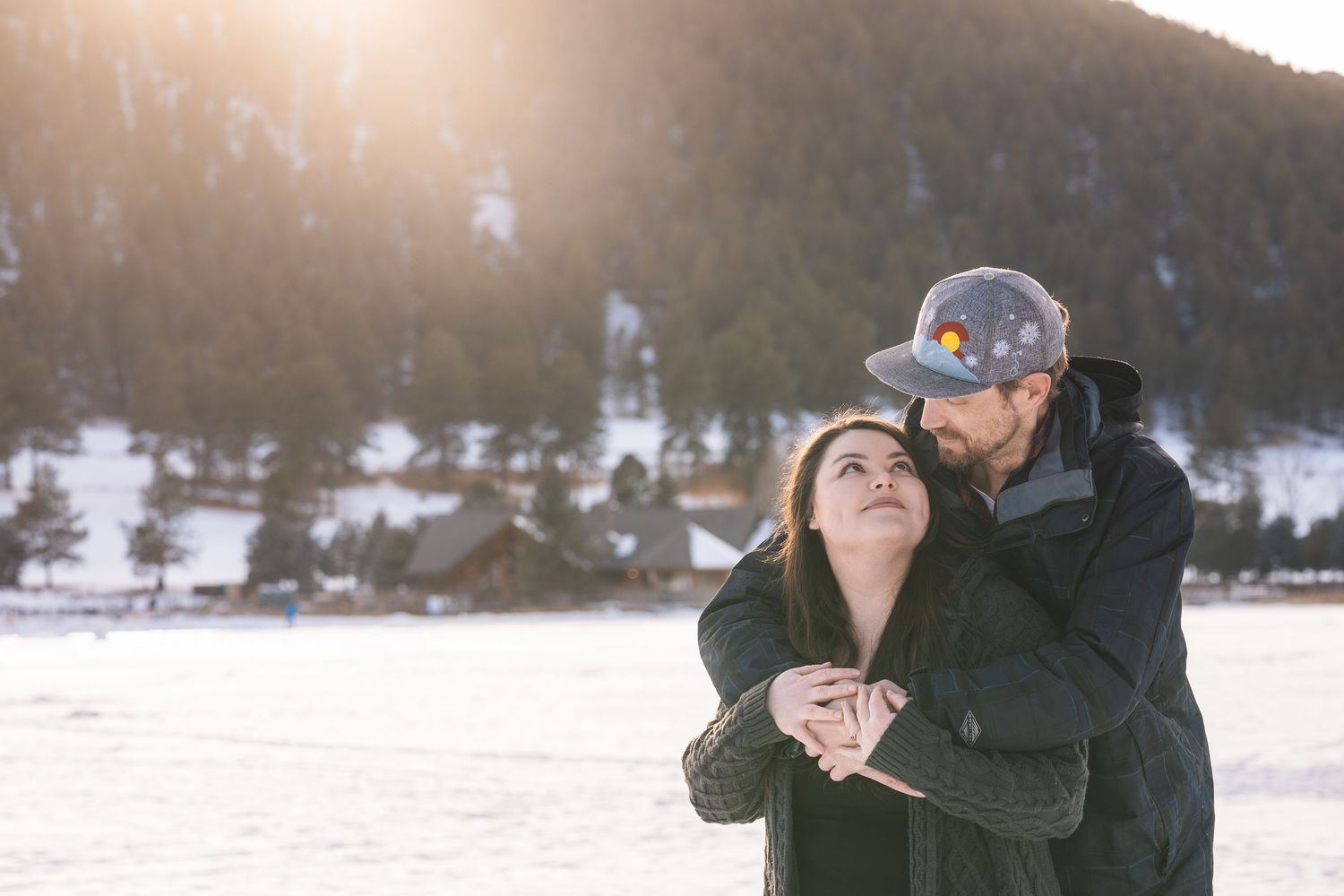 A winter scene with a couple sharing a loving moment in the snow with mountains behind them.