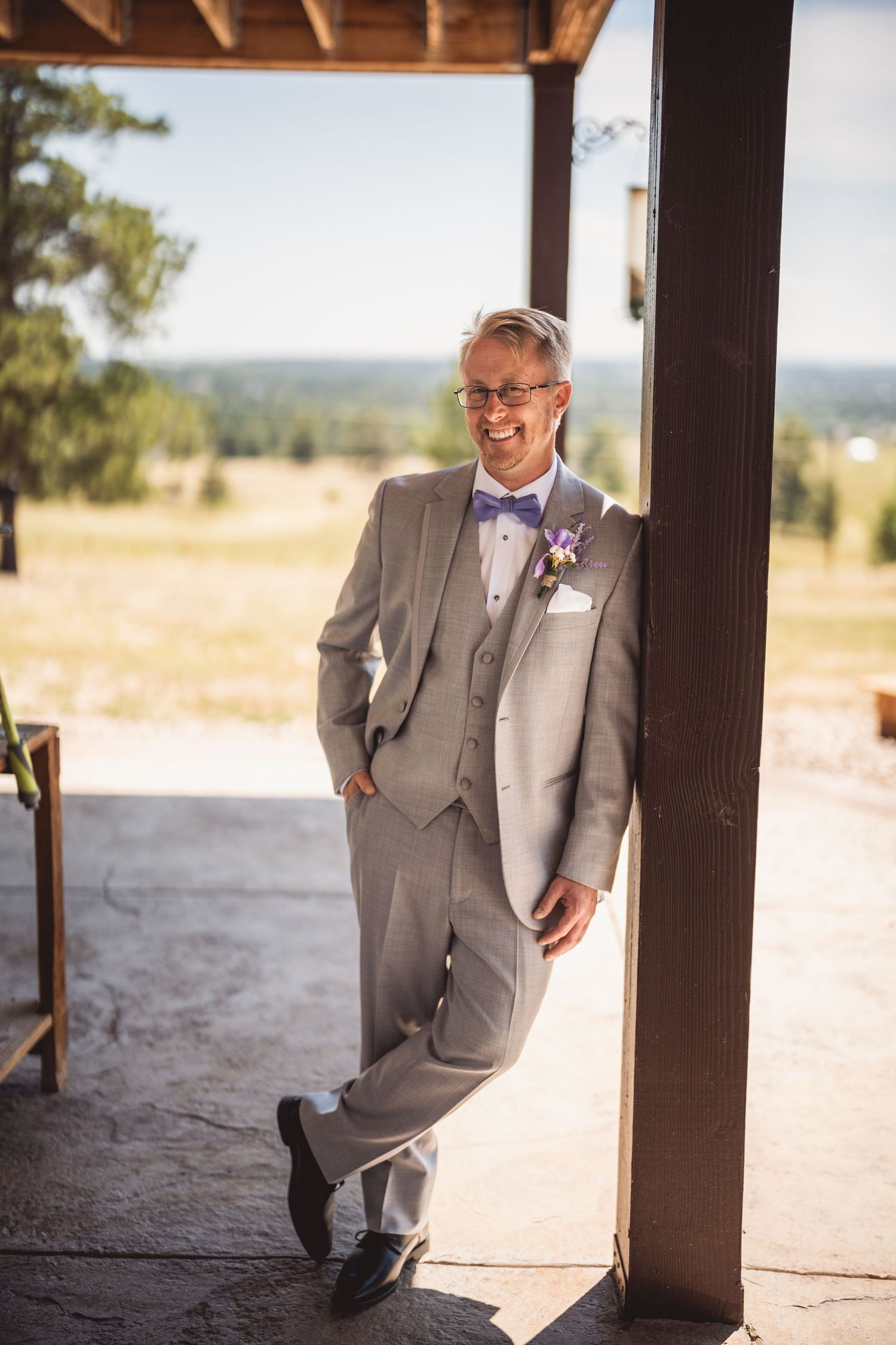 A groom in a grey suit poses casually against a rustic wooden backdrop.