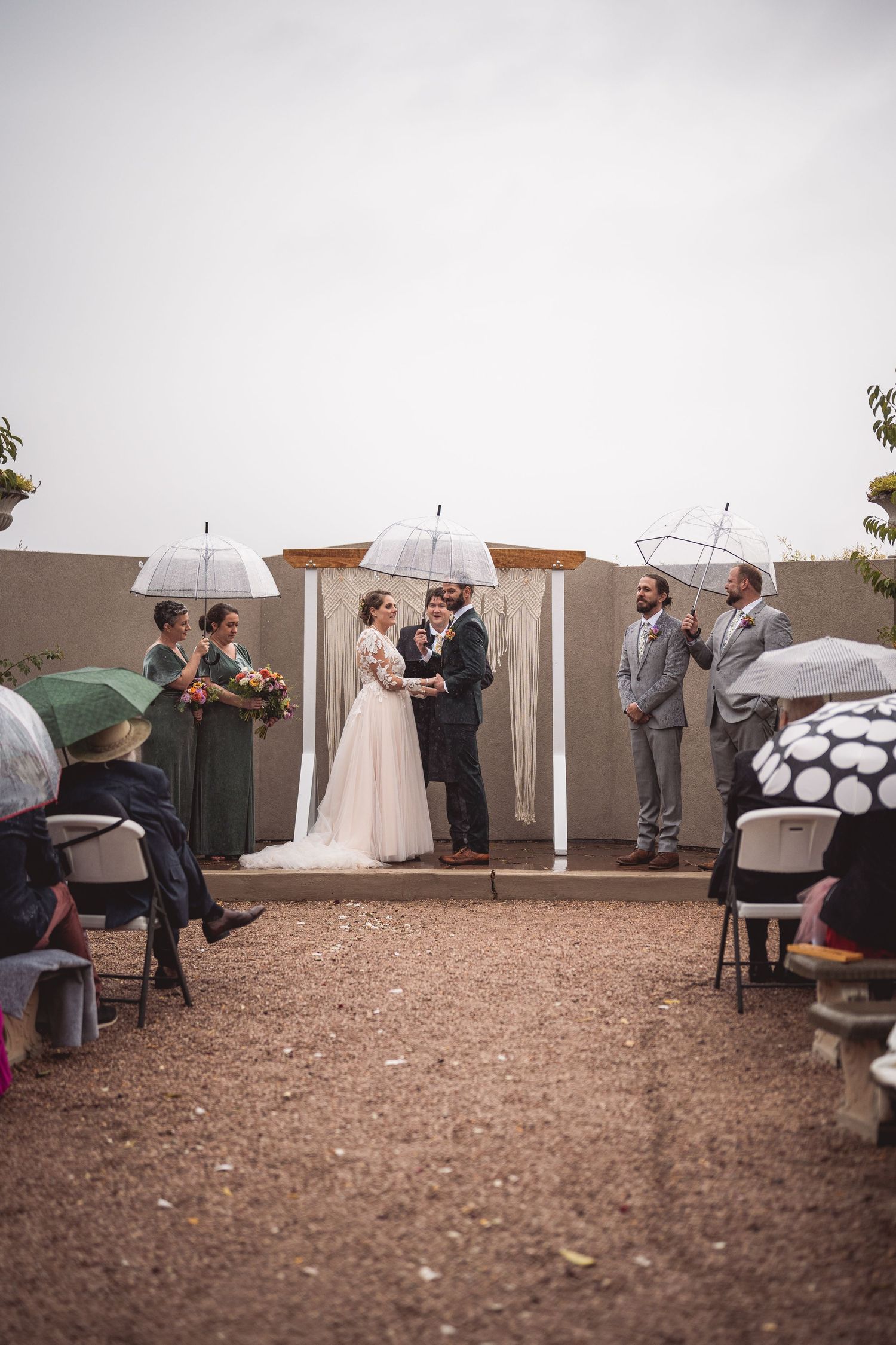 Wedding ceremony panorama with guests holding white umbrellas on gravel ground.