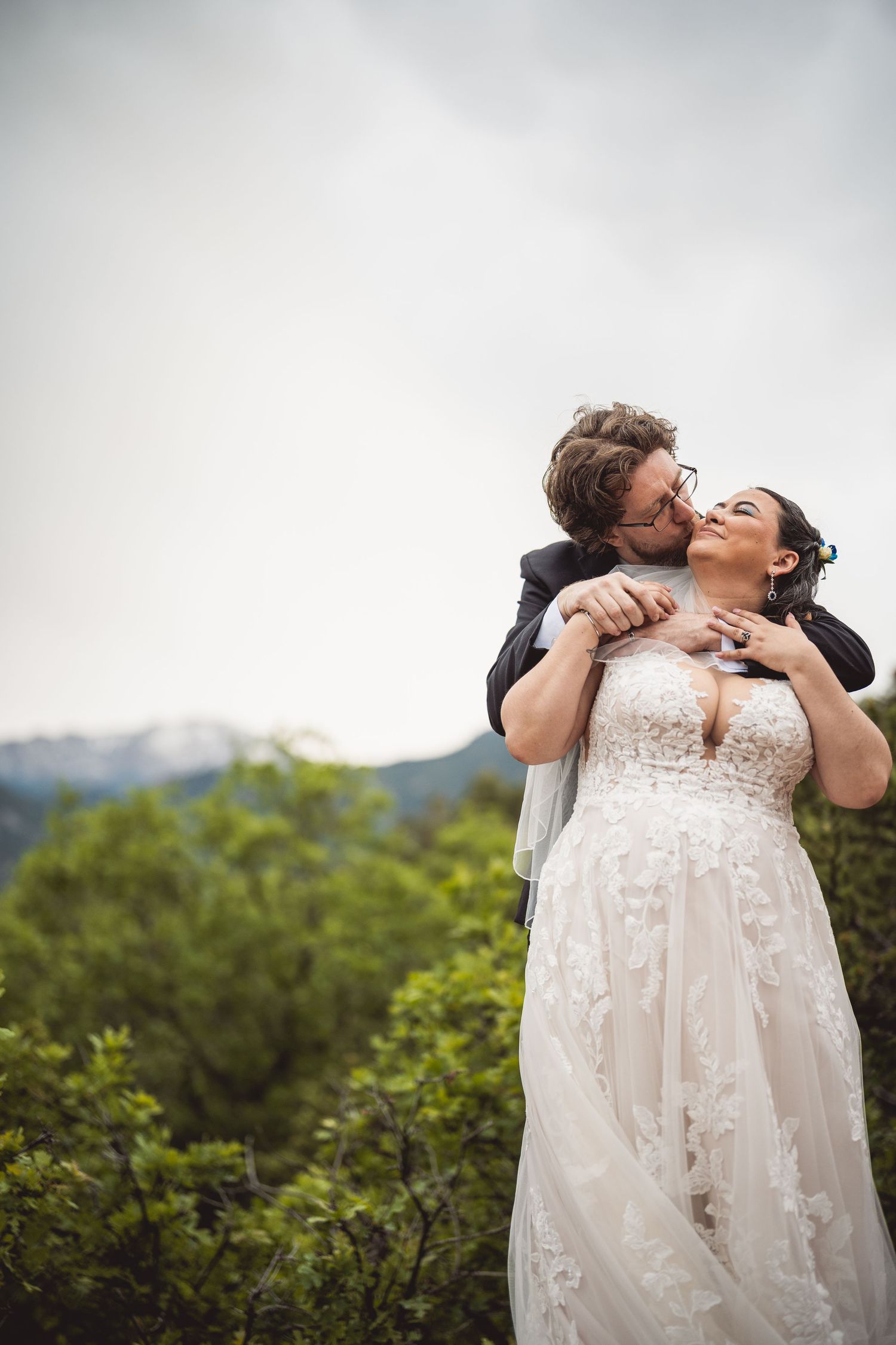 Wedding couple shares a romantic moment outdoors with mountains and greenery in the background.