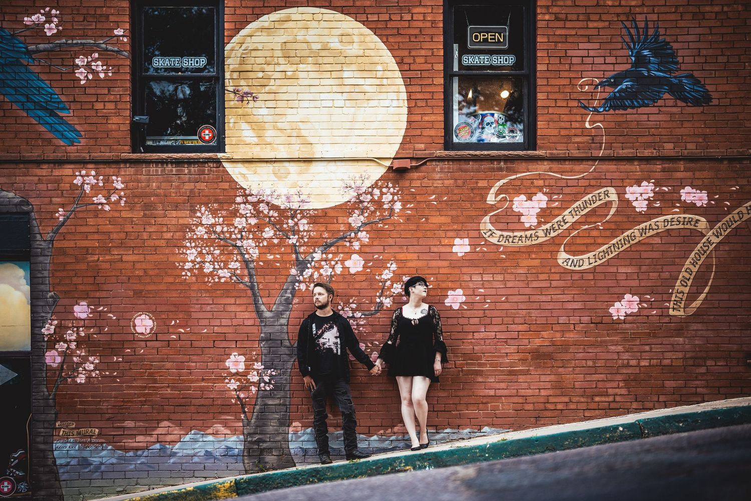 A couple poses against a brick wall with artistic moon and bird murals.