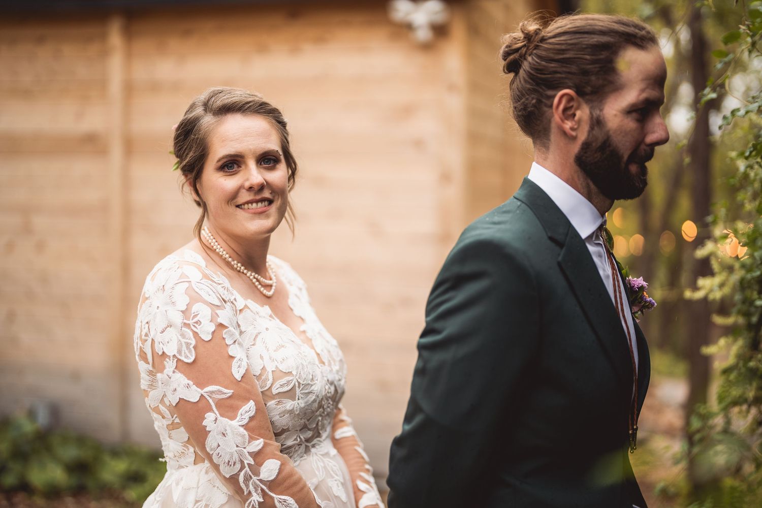 Bride in lace wedding dress smiles at groom in dark green suit during outdoor wedding ceremony.