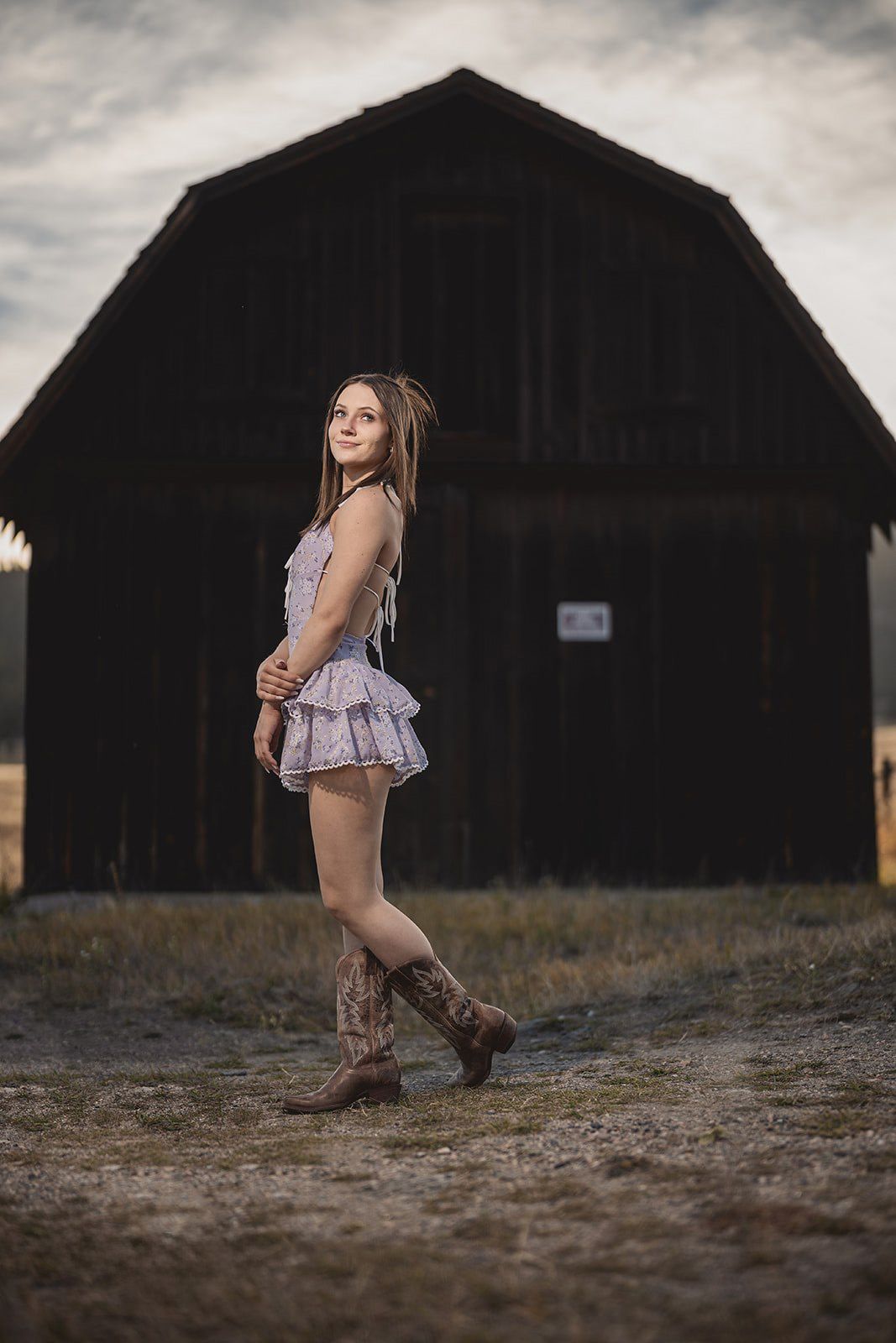 Young woman in ruffled dress and cowboy boots posing before dark weathered barn.