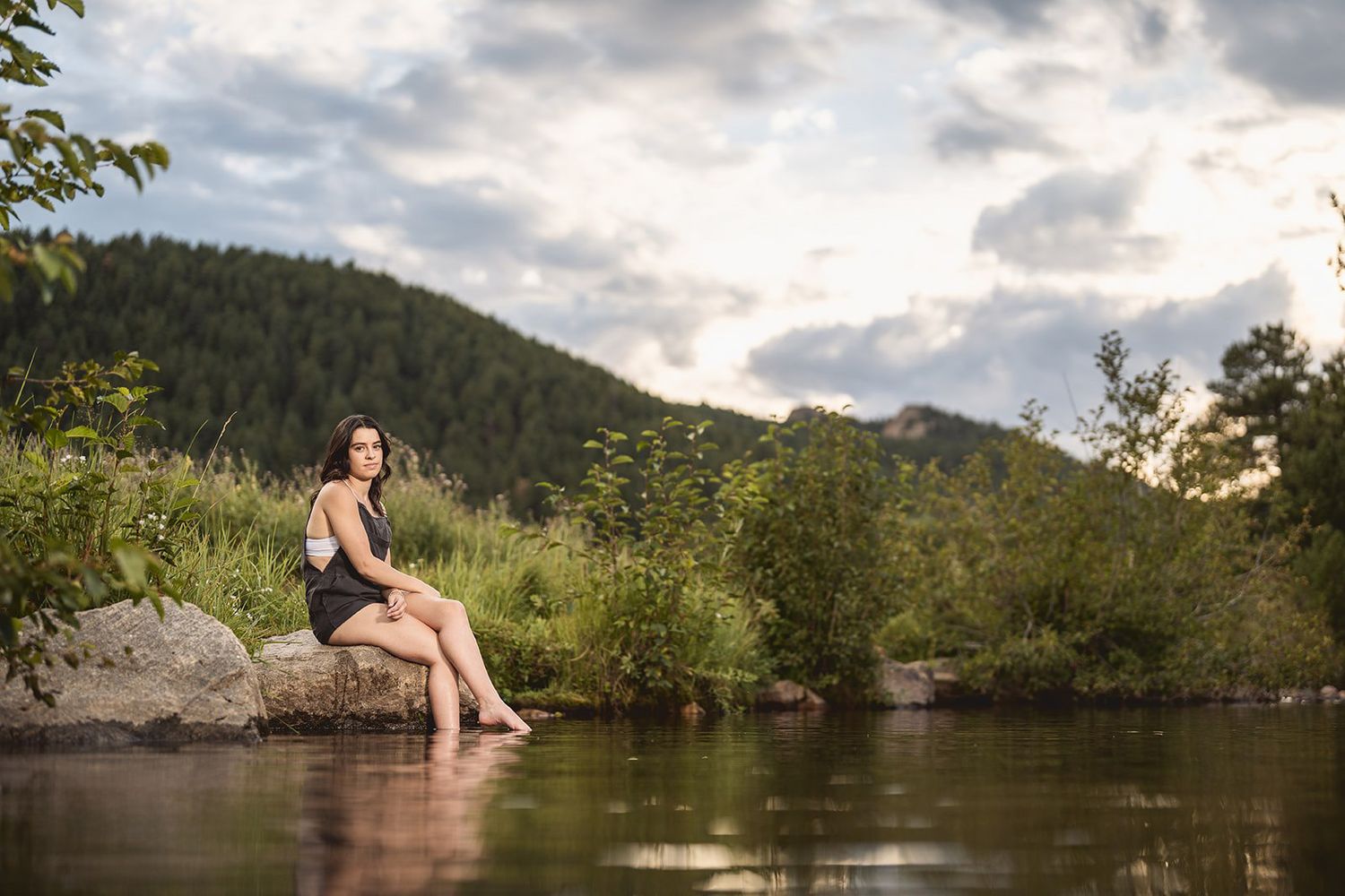 Young woman in black swimsuit sitting on a rock by a peaceful river with lush green hills and dramatic cloudy sky.