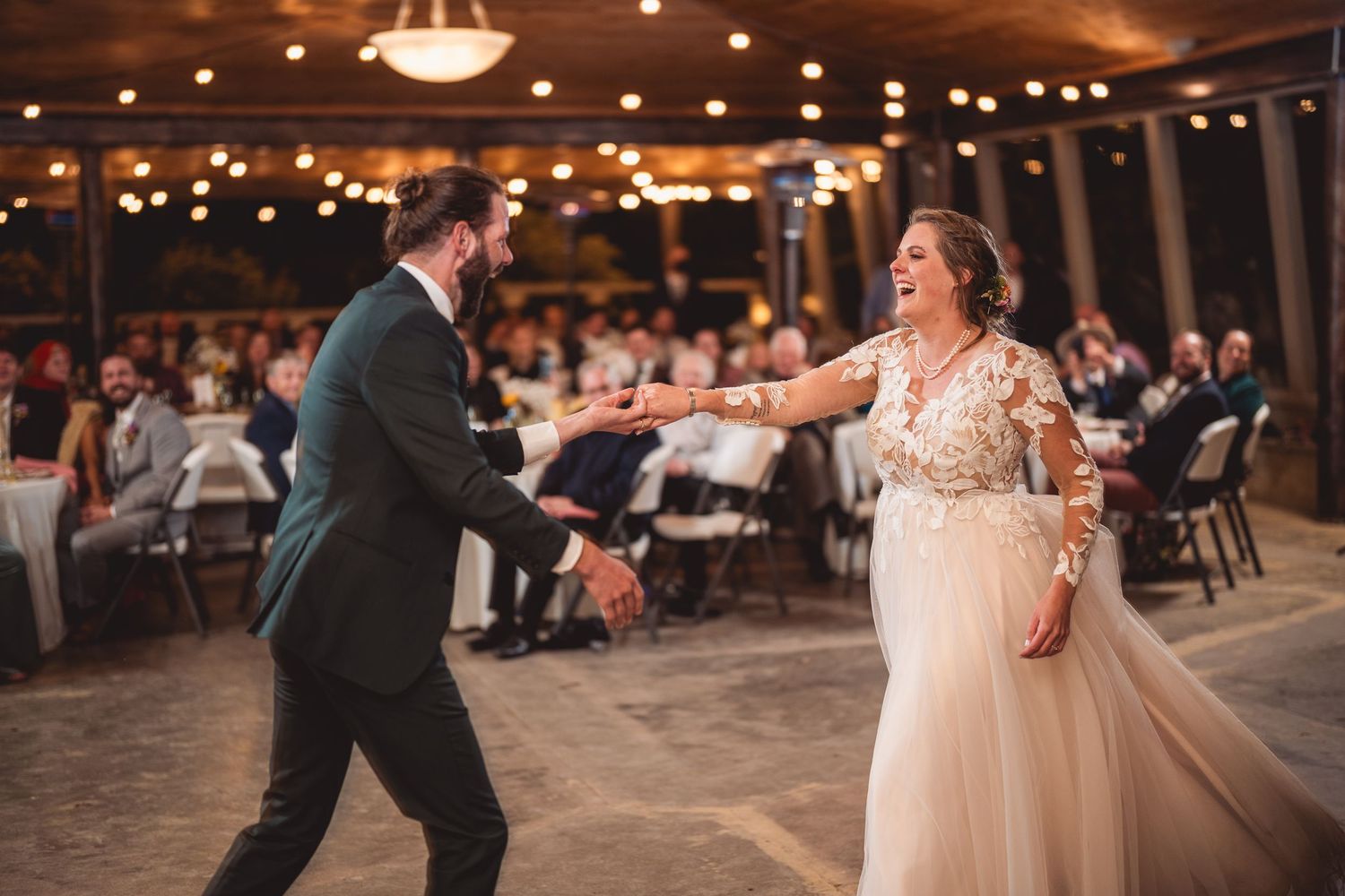 Couple dances together in an elegant rustic venue with string lights and white table settings.