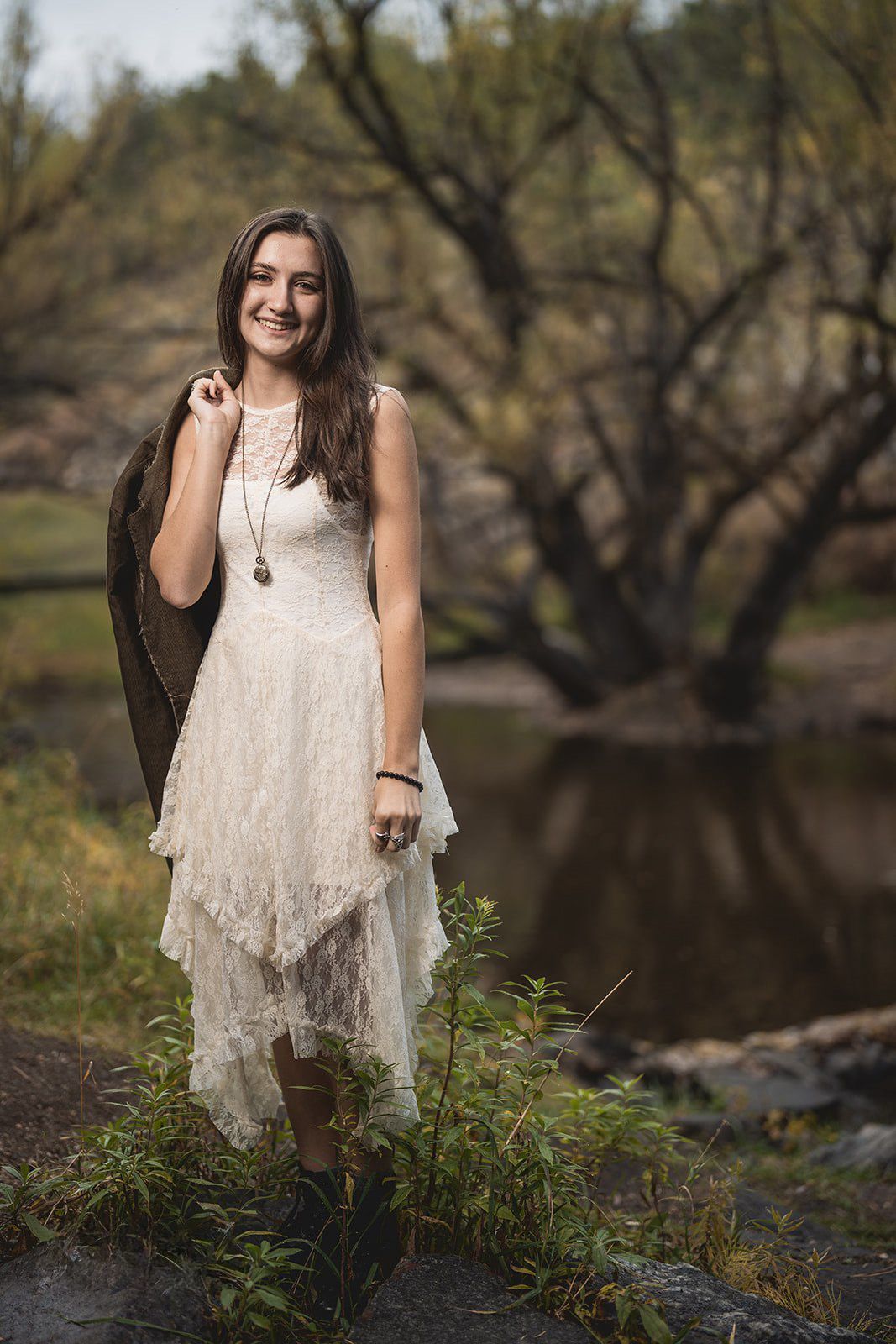 Nature portrait session by a river with white lace dress and autumn trees in background.