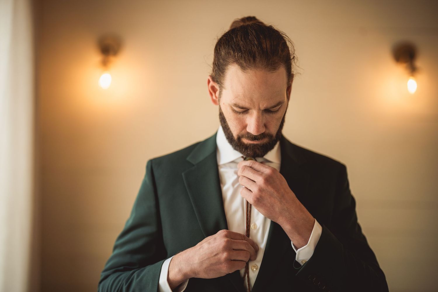 Person in dark green suit adjusting tie in dimly lit room.