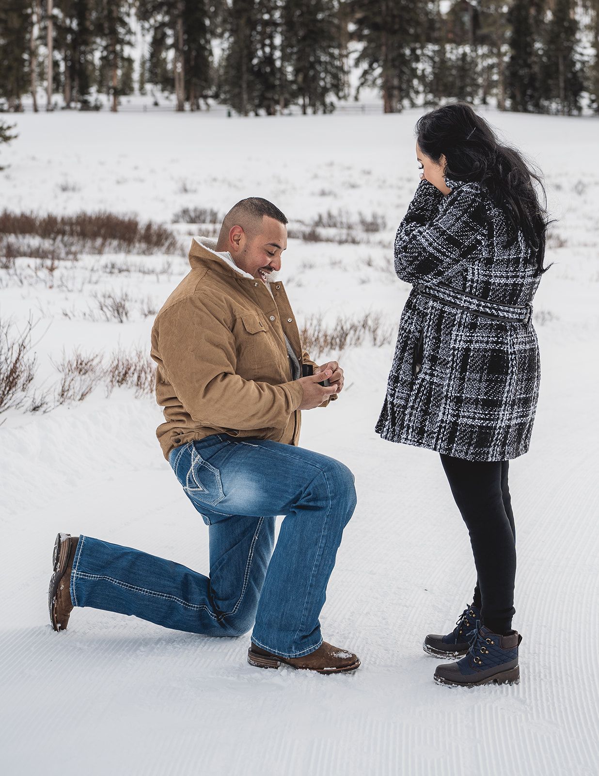 Man proposes on one knee to woman in snowy winter landscape, both wearing warm coats outdoors.