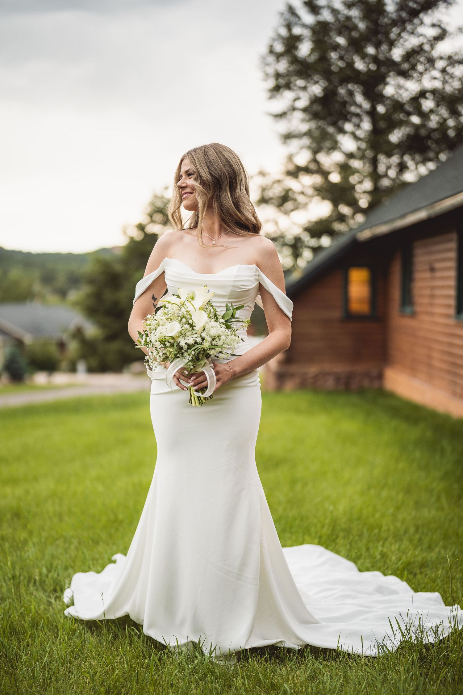 Smiling bride in off-shoulder white gown holding floral bouquet standing on green lawn near rustic wooden building.