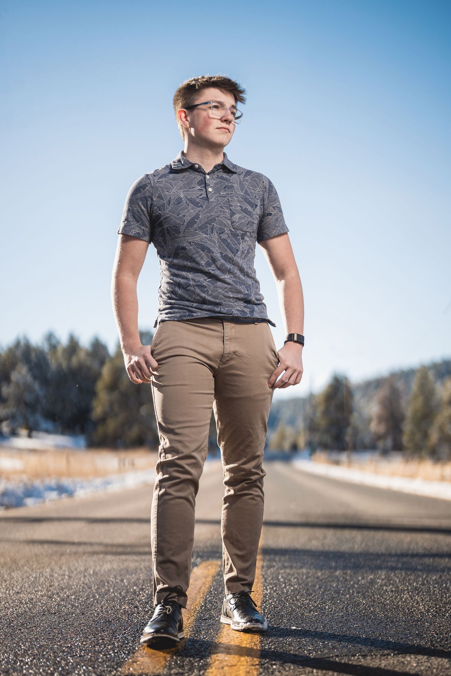 Young man in glasses and khaki pants stands confidently on an empty road with snowy trees in background.