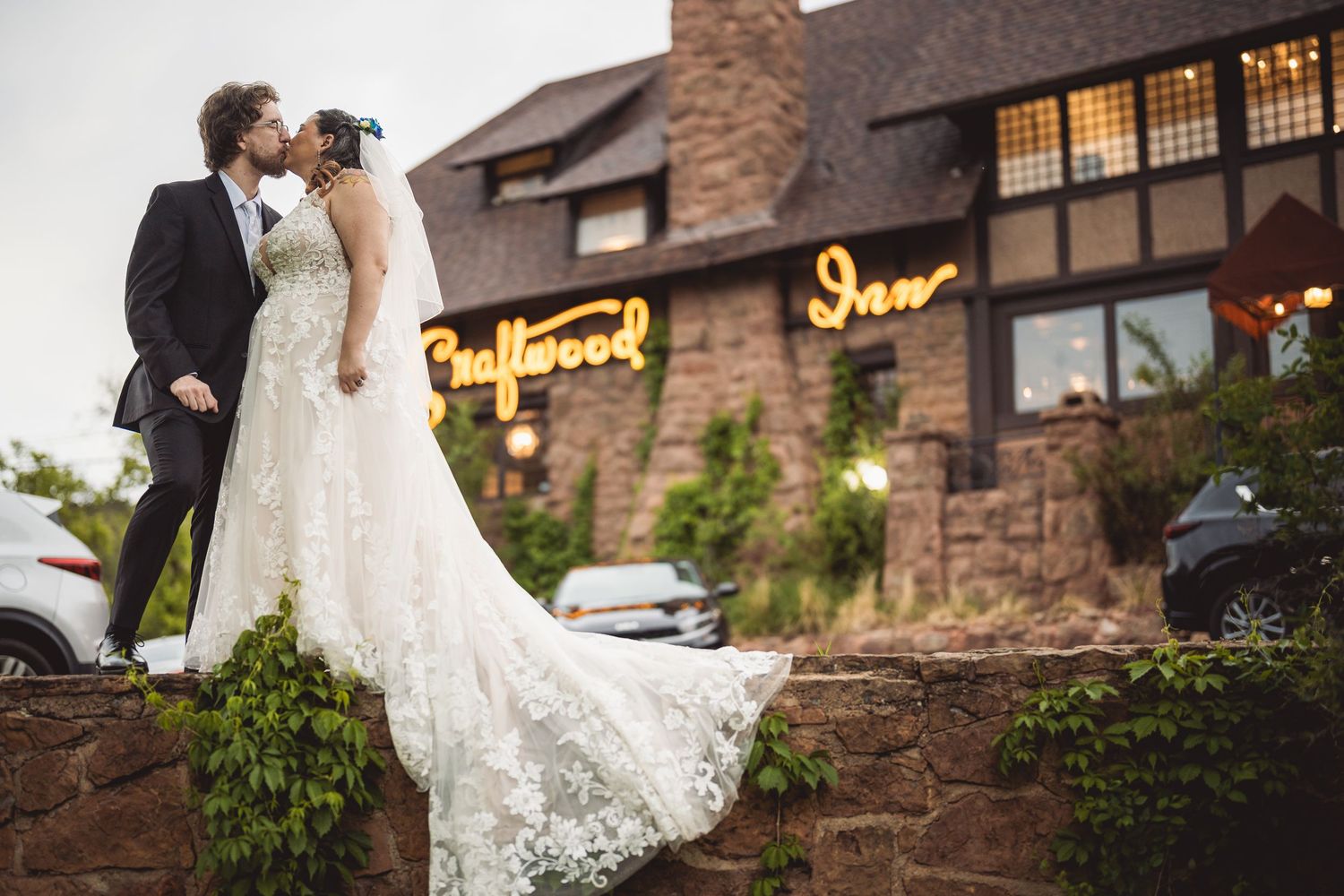 Bride and groom kiss on stone wall before the Craftwood Inn at dusk.