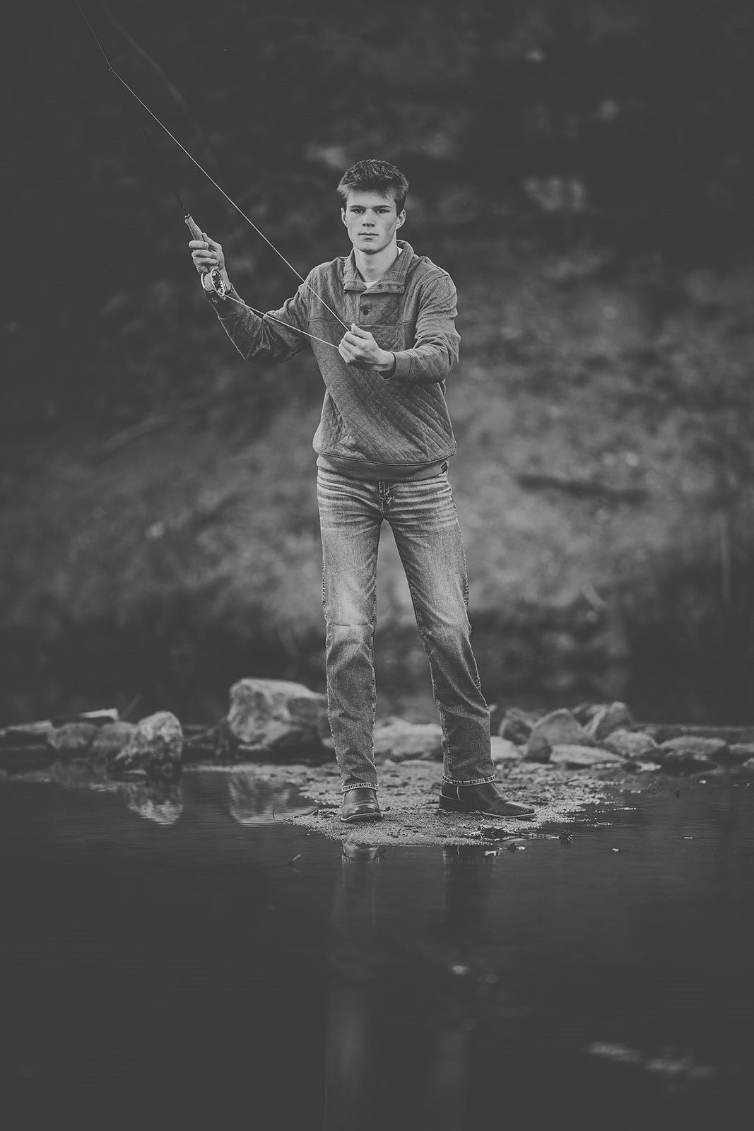 Black and white photo of a young man fly fishing in a rocky stream, holding a fishing rod.