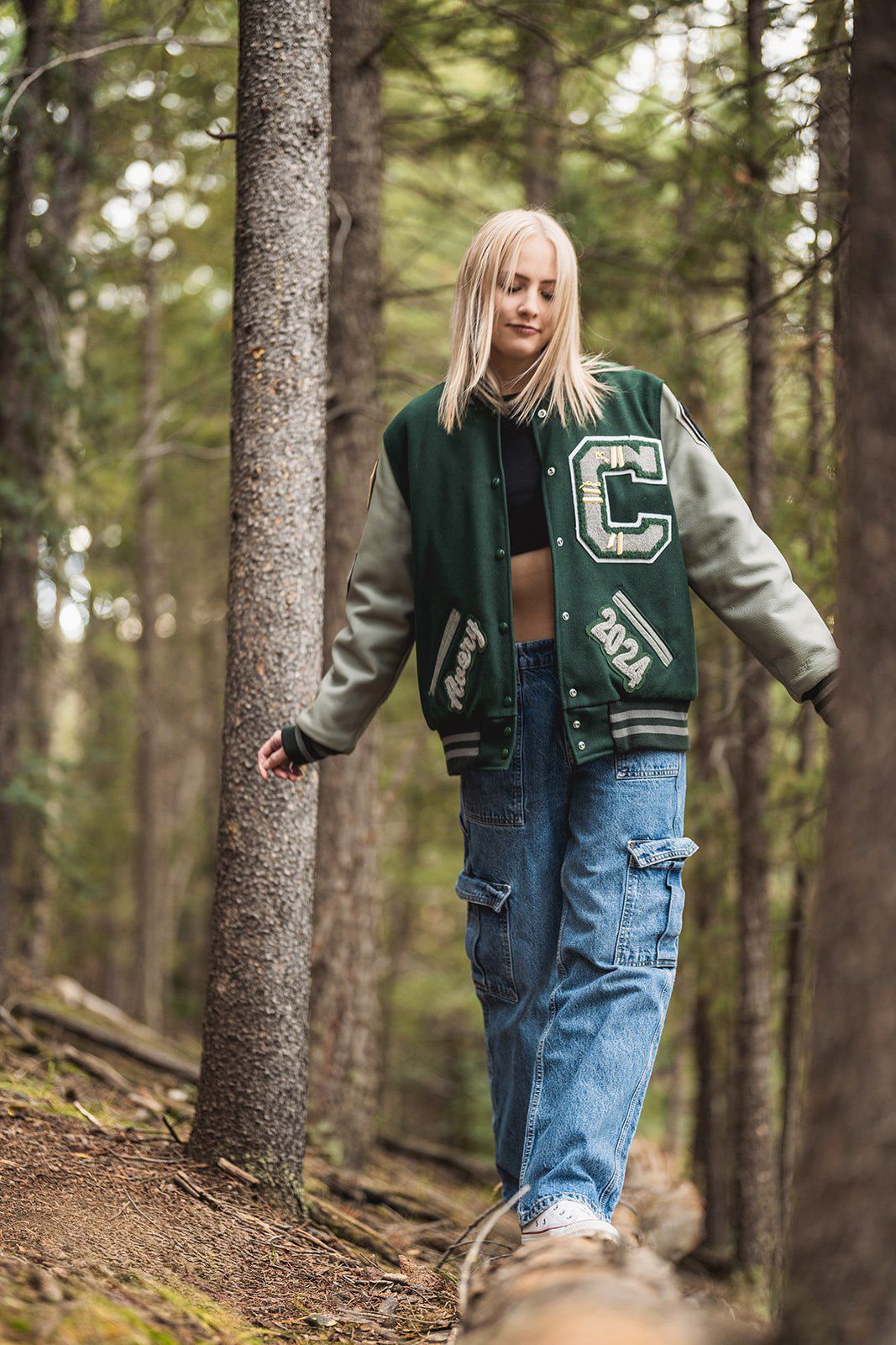 Blonde teen in green varsity jacket and ripped jeans walking through pine forest on log.