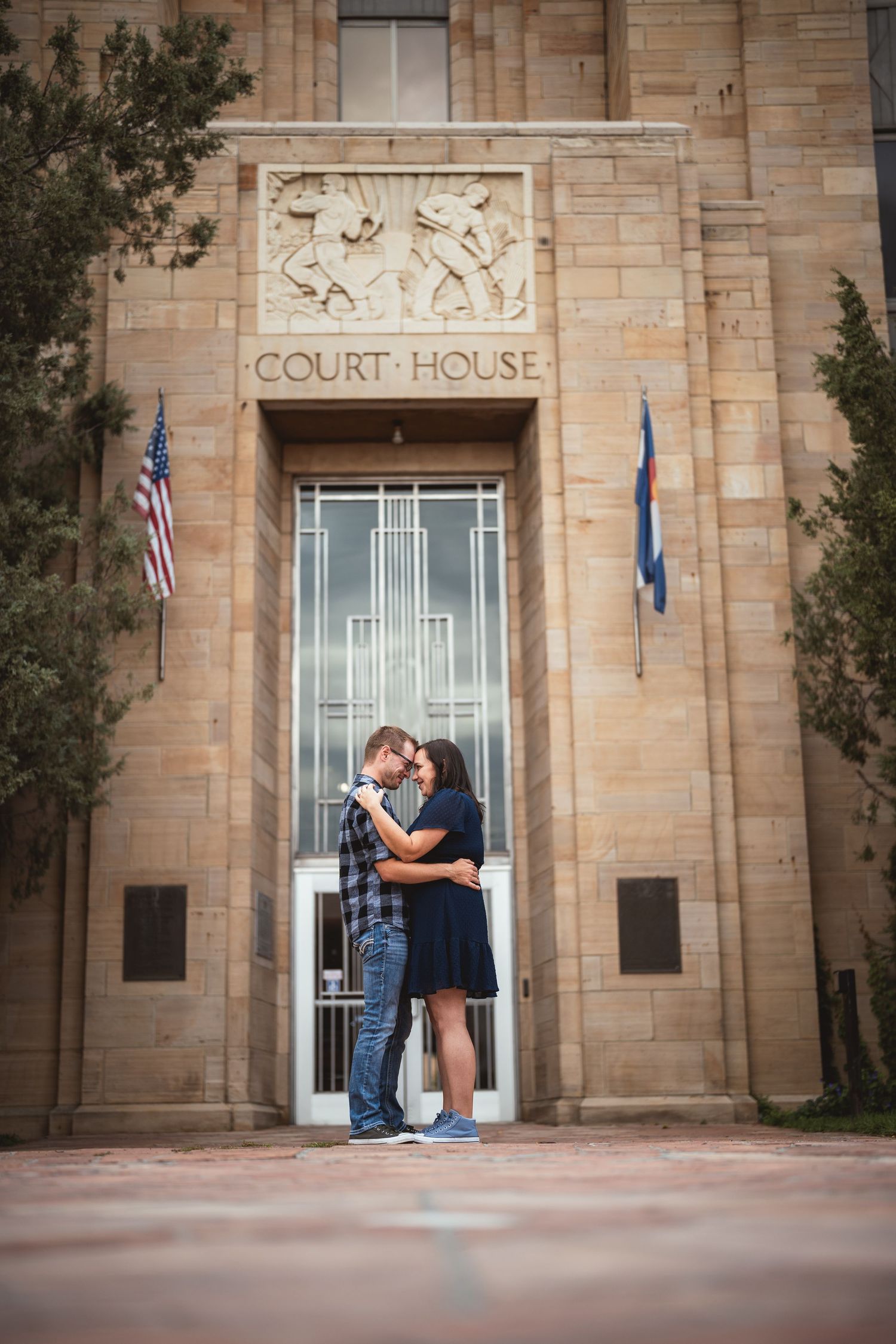 Couple embracing romantically in front of a historic courthouse building with American and state flags.