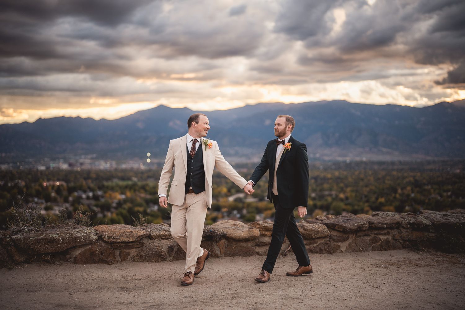 Two grooms hold hands walking at sunset with mountain skyline backdrop on their wedding day.