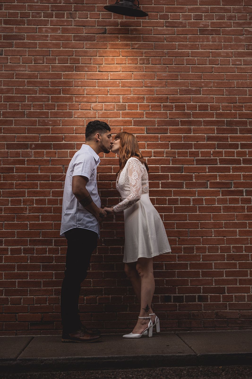 A romantic couple shares a kiss against a red brick wall under warm lighting, woman in white lace dress and heels.