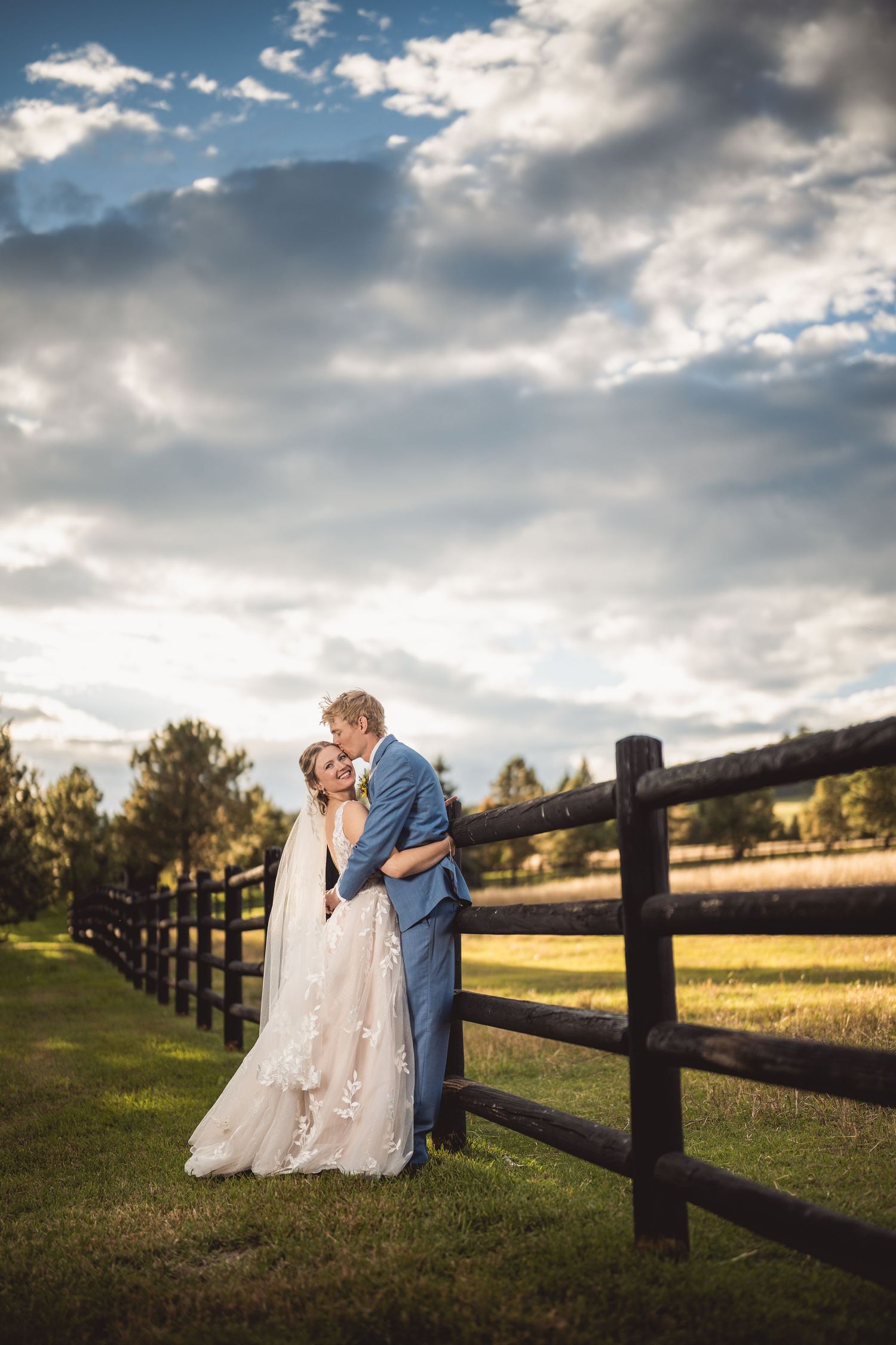 Bride and groom embrace by rustic wooden fence in lush green field under dramatic cloudy sky at outdoor wedding.
