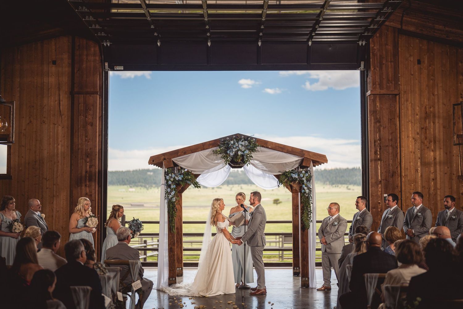 Couple exchanges vows under floral arch in rustic barn wedding venue with scenic countryside views.