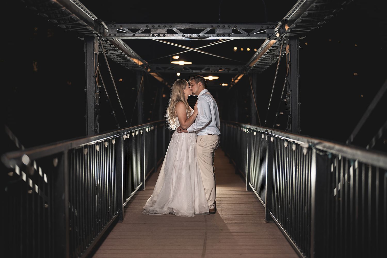 Bride and groom sharing a romantic kiss on a dimly lit metal bridge at night during their wedding celebration.