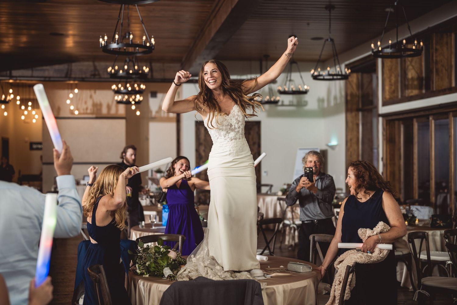 Bride in white gown dancing on table at wedding reception with guests celebrating around her.