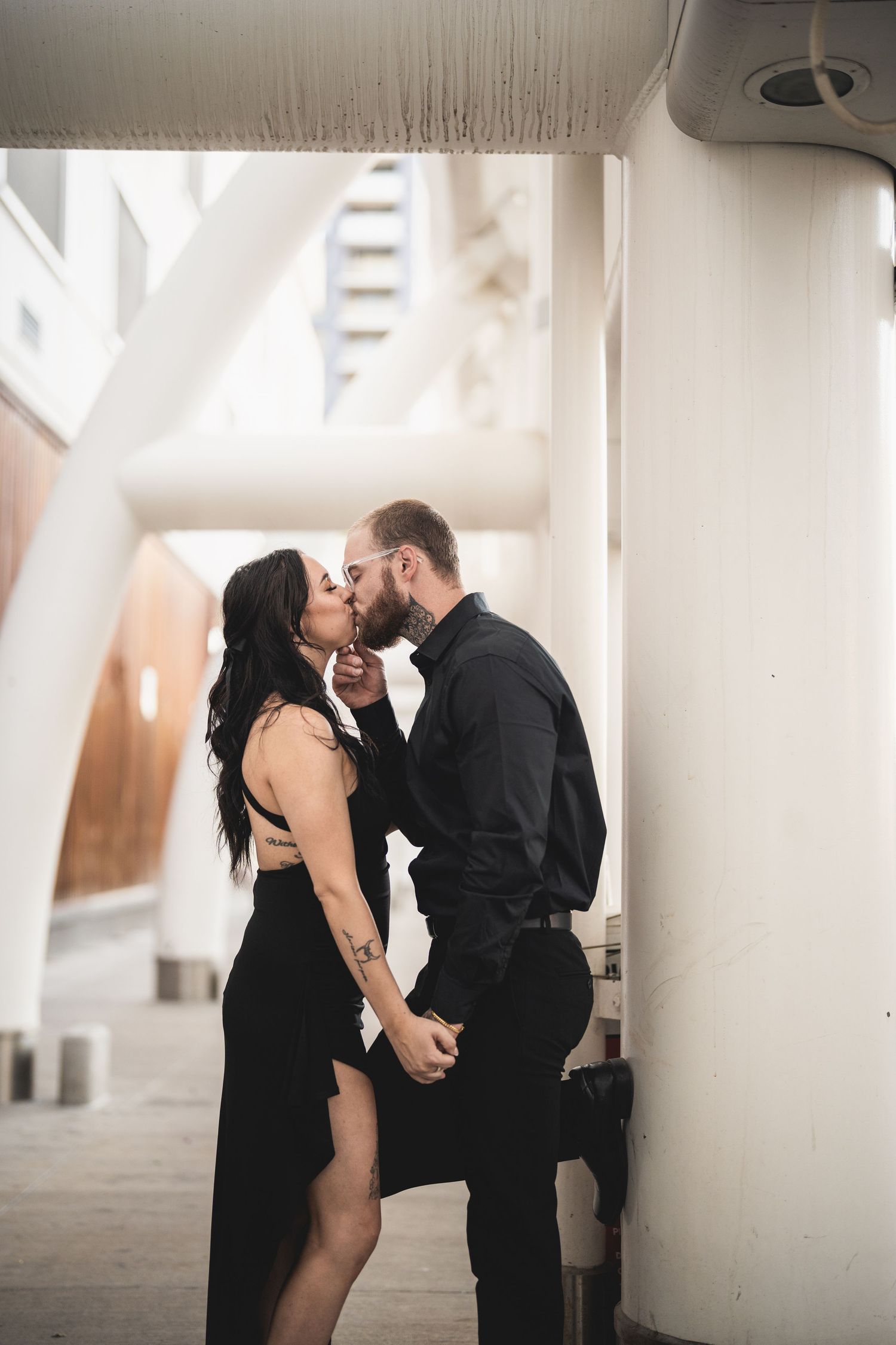 Couple in black outfits kissing between white pillars in an urban setting, woman in backless dress holding hands with man.