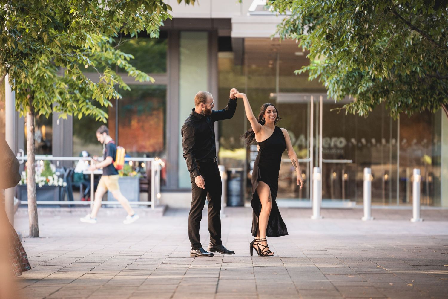 Couple dancing tango outdoors in black attire on urban plaza surrounded by trees and modern buildings.