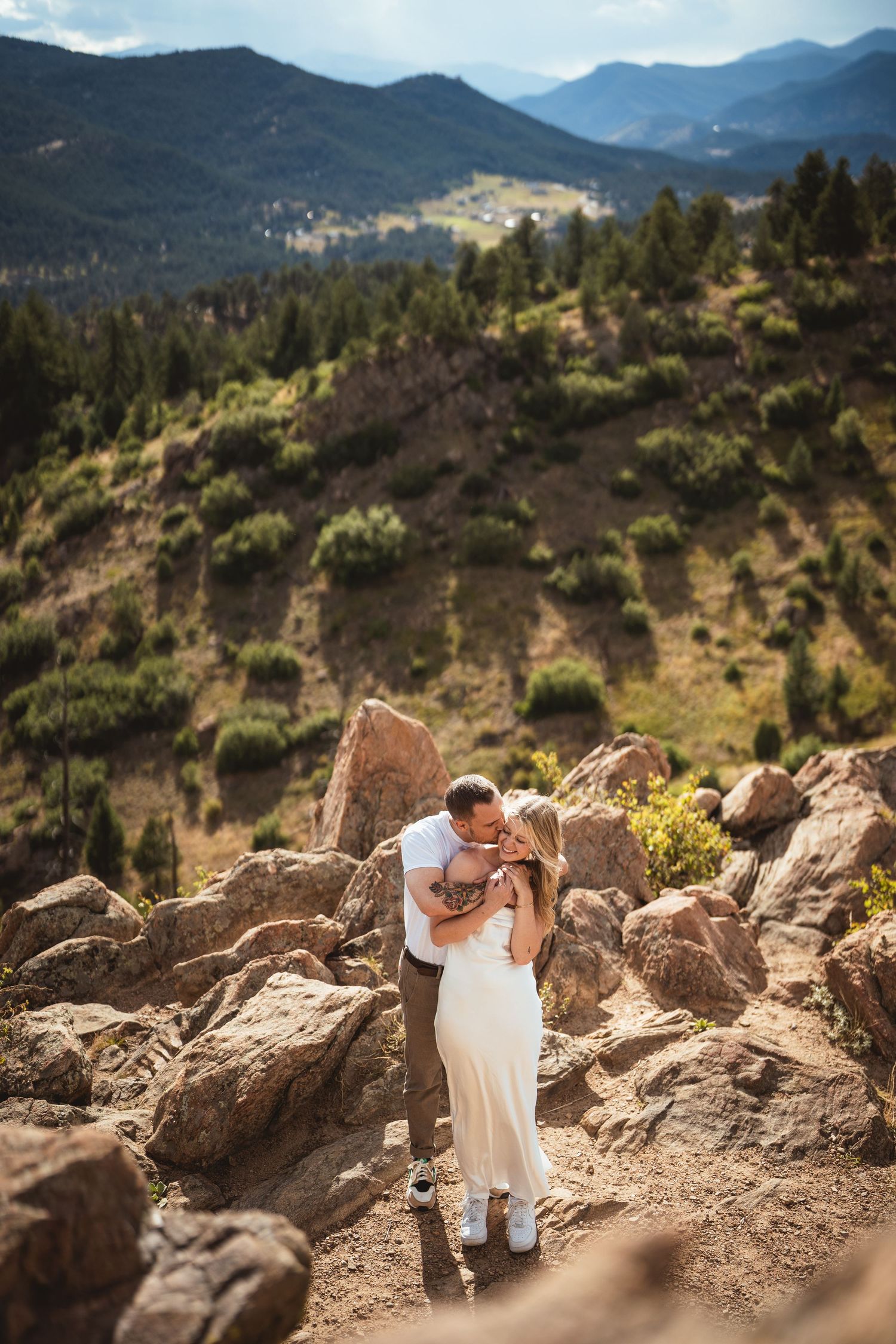 Couple embracing on rocky mountain trail with lush green pine forest and scenic Colorado mountain range in background.