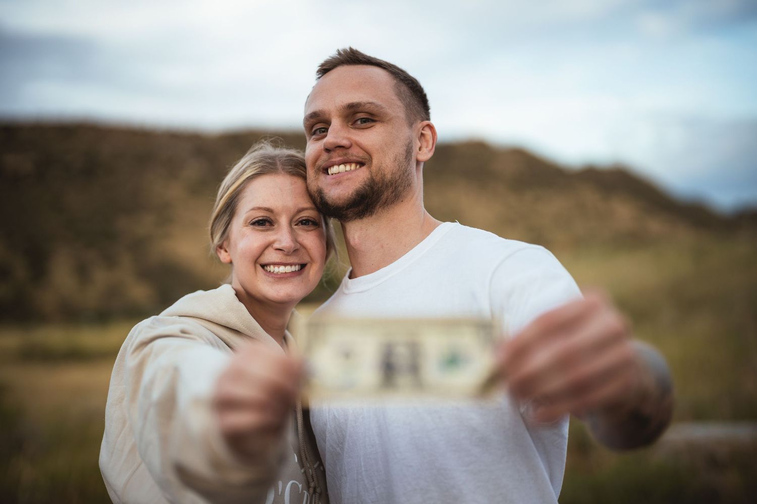 Happy couple smiling and holding a dollar bill outdoors with green hills in the background.