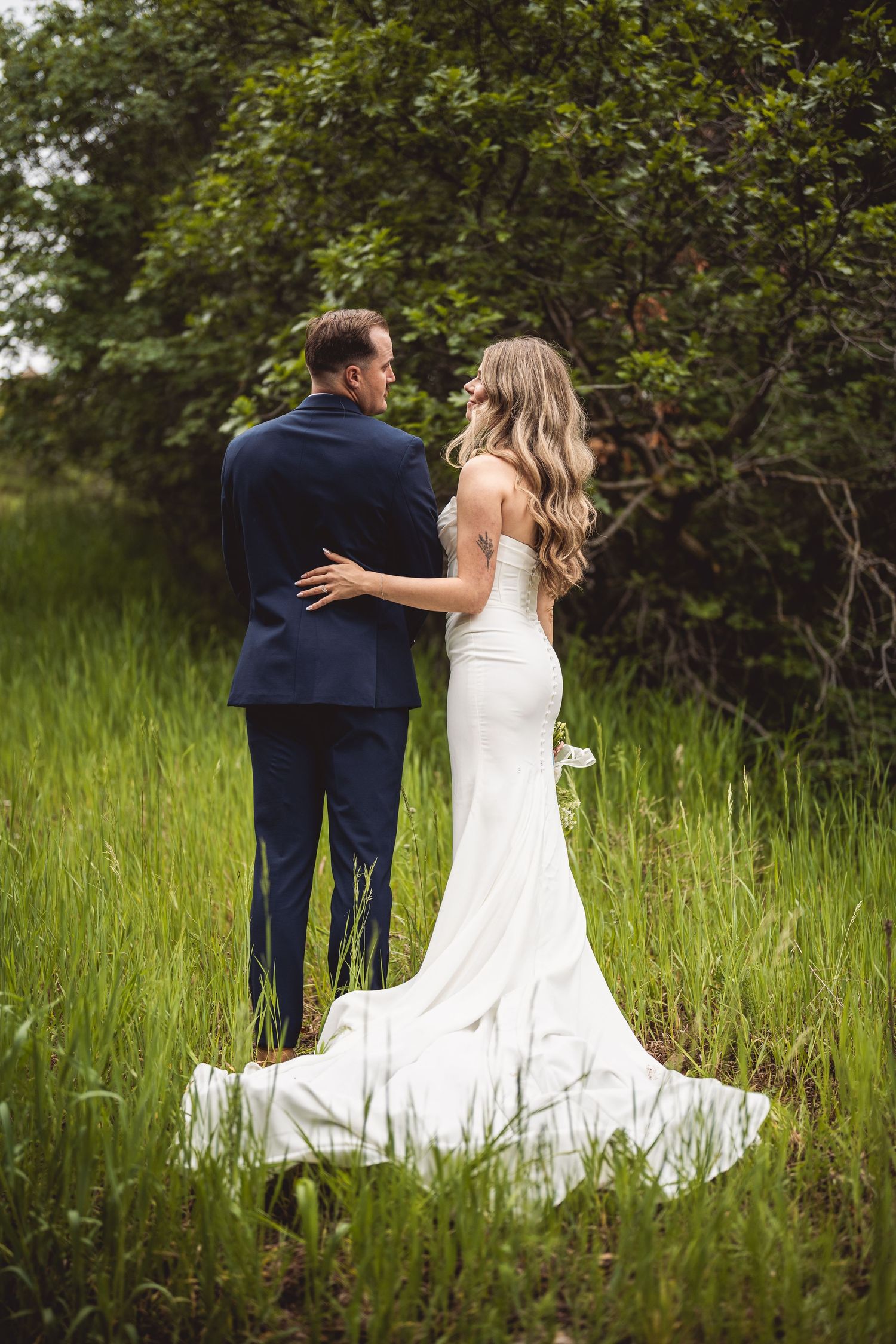 A newlywed couple in blue suit and white dress standing together in tall grass with trees in the background.