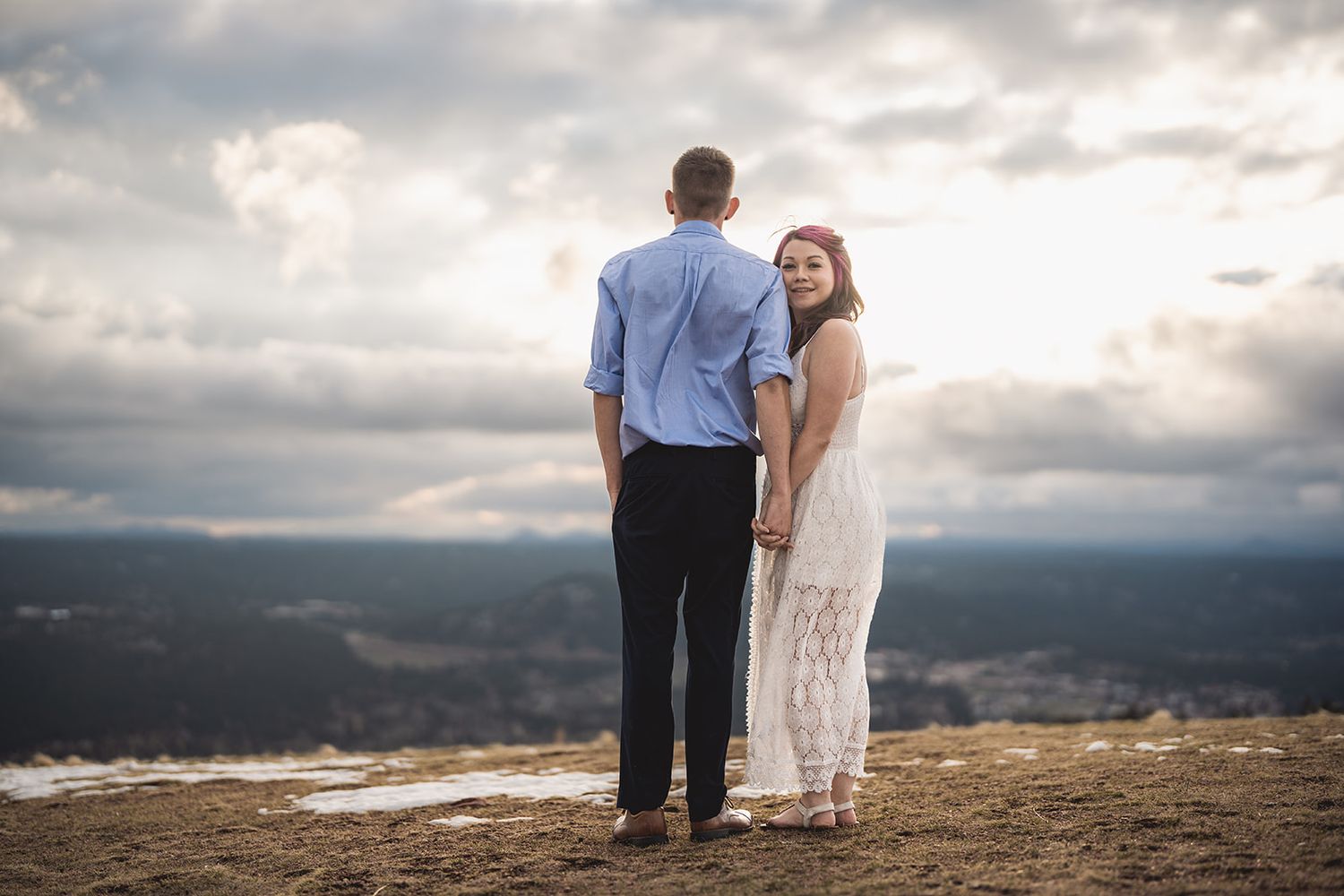 Couple embracing on mountain overlook with dramatic cloudy sky and valley views.