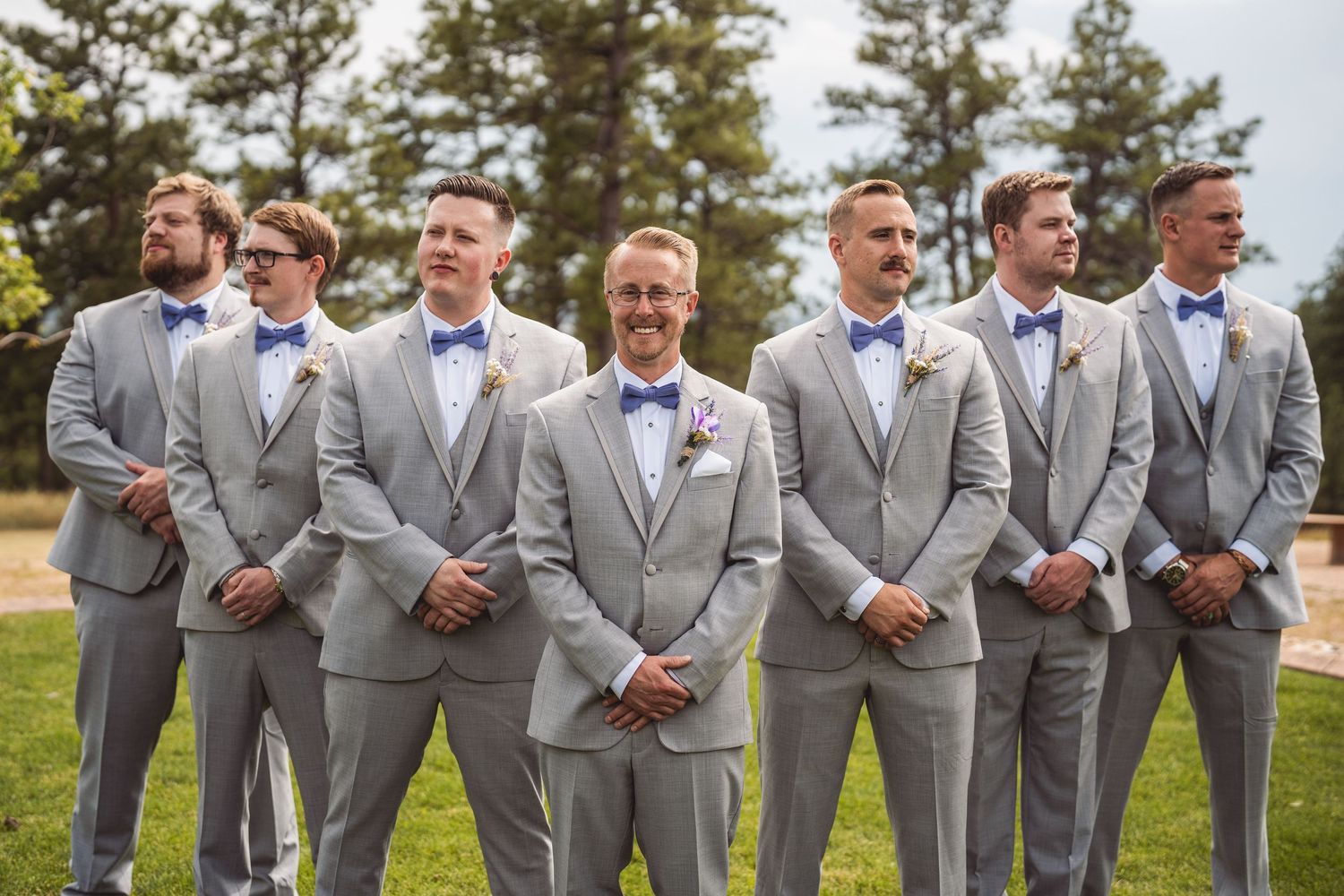 Wedding party in gray suits stands together outdoors for candid group photos.