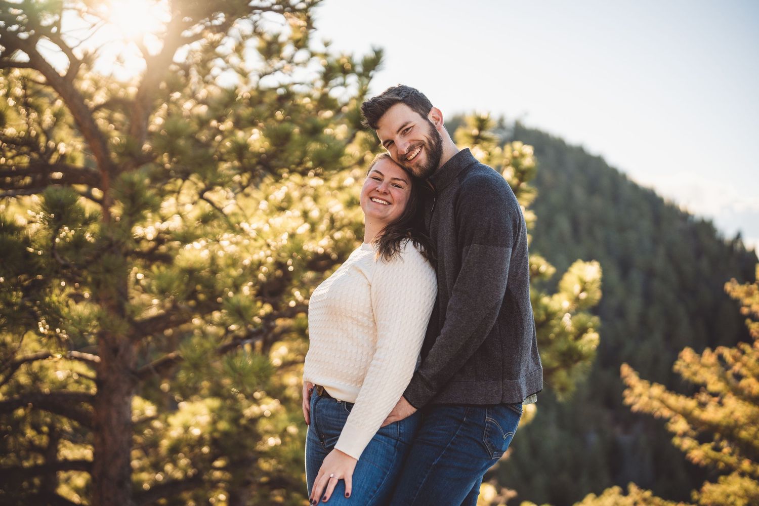 A tender embrace against a backdrop of sunlit mountain scenery and pine trees.