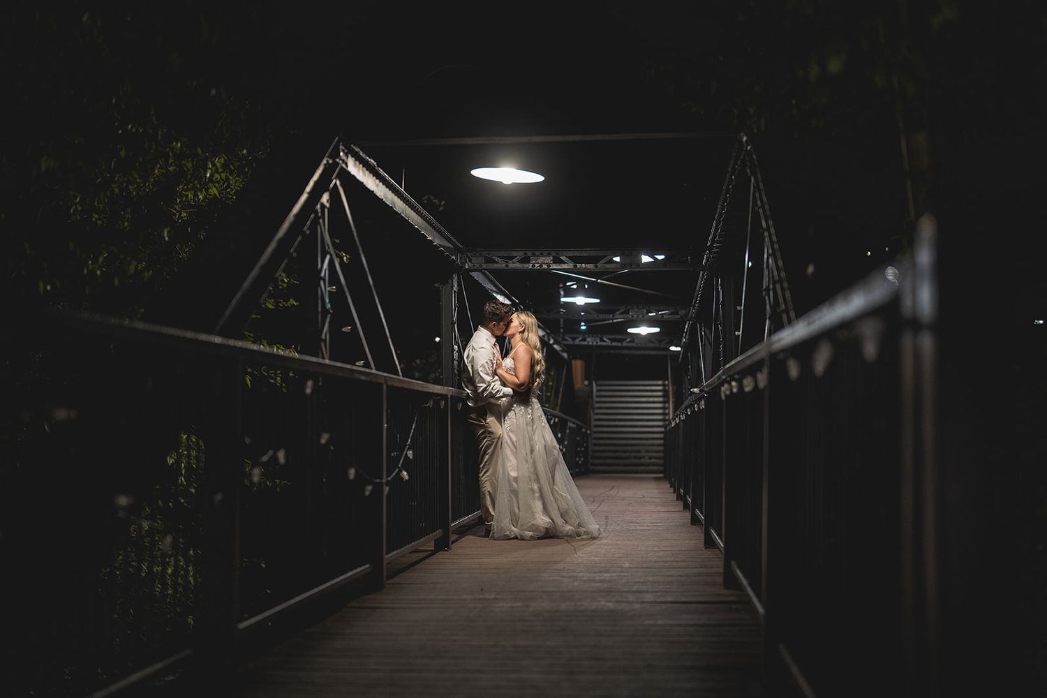 Couple sharing a romantic kiss on a dimly lit bridge at night in wedding attire.