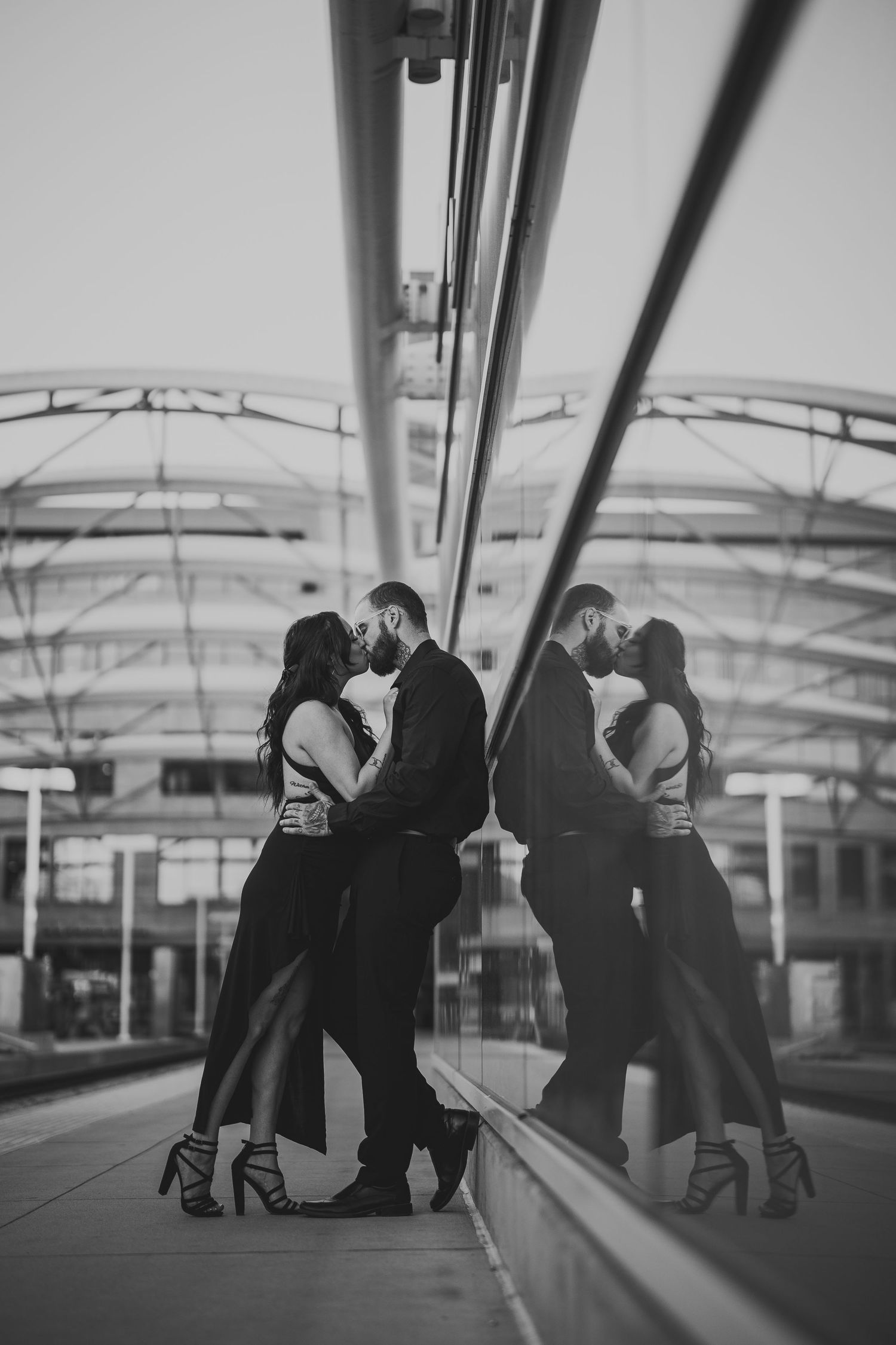 Black and white photo of a couple kissing beside a glass wall reflection in a modern building.