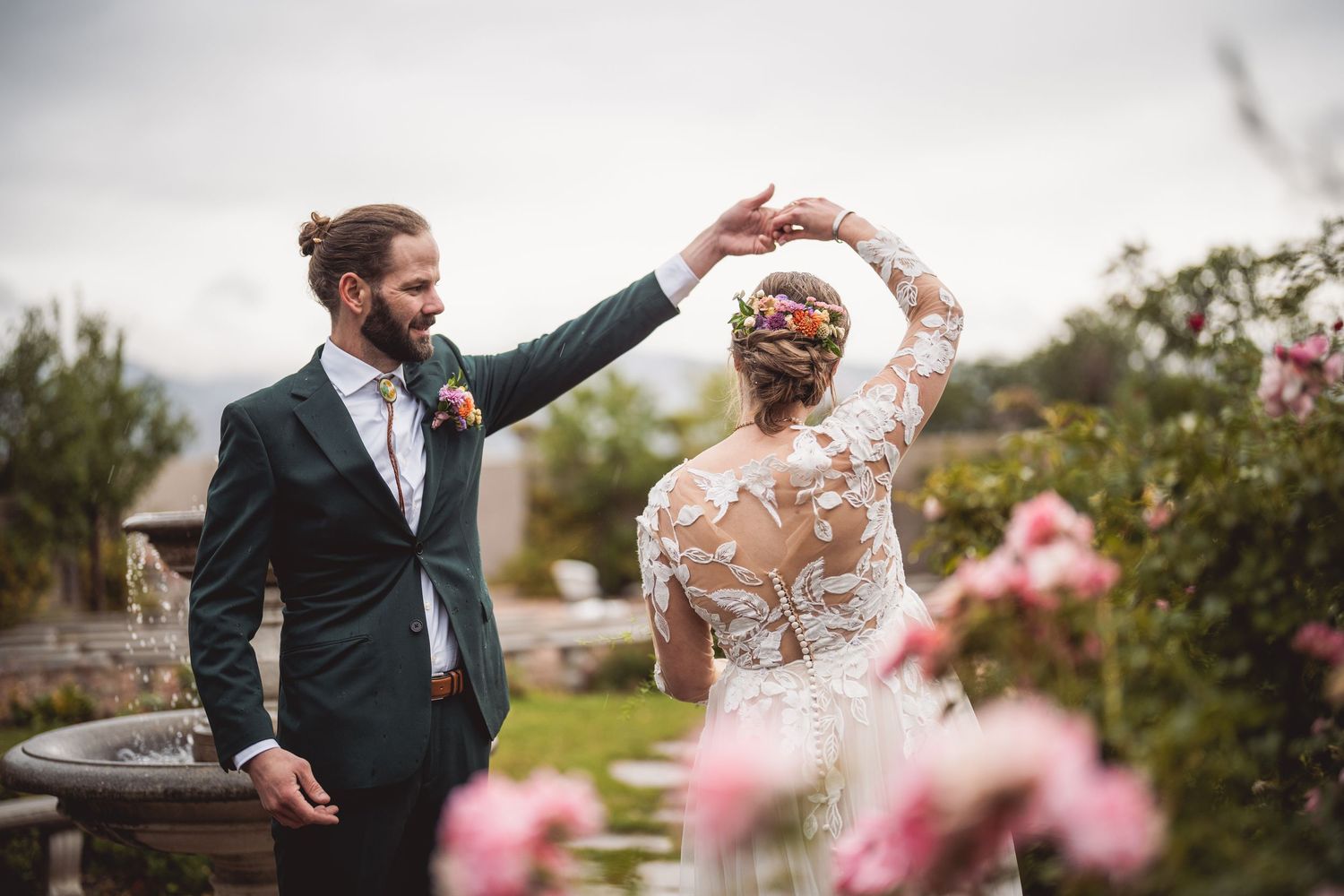 Dancing wedding couple twirling in garden surrounded by pink flowers and stone elements.