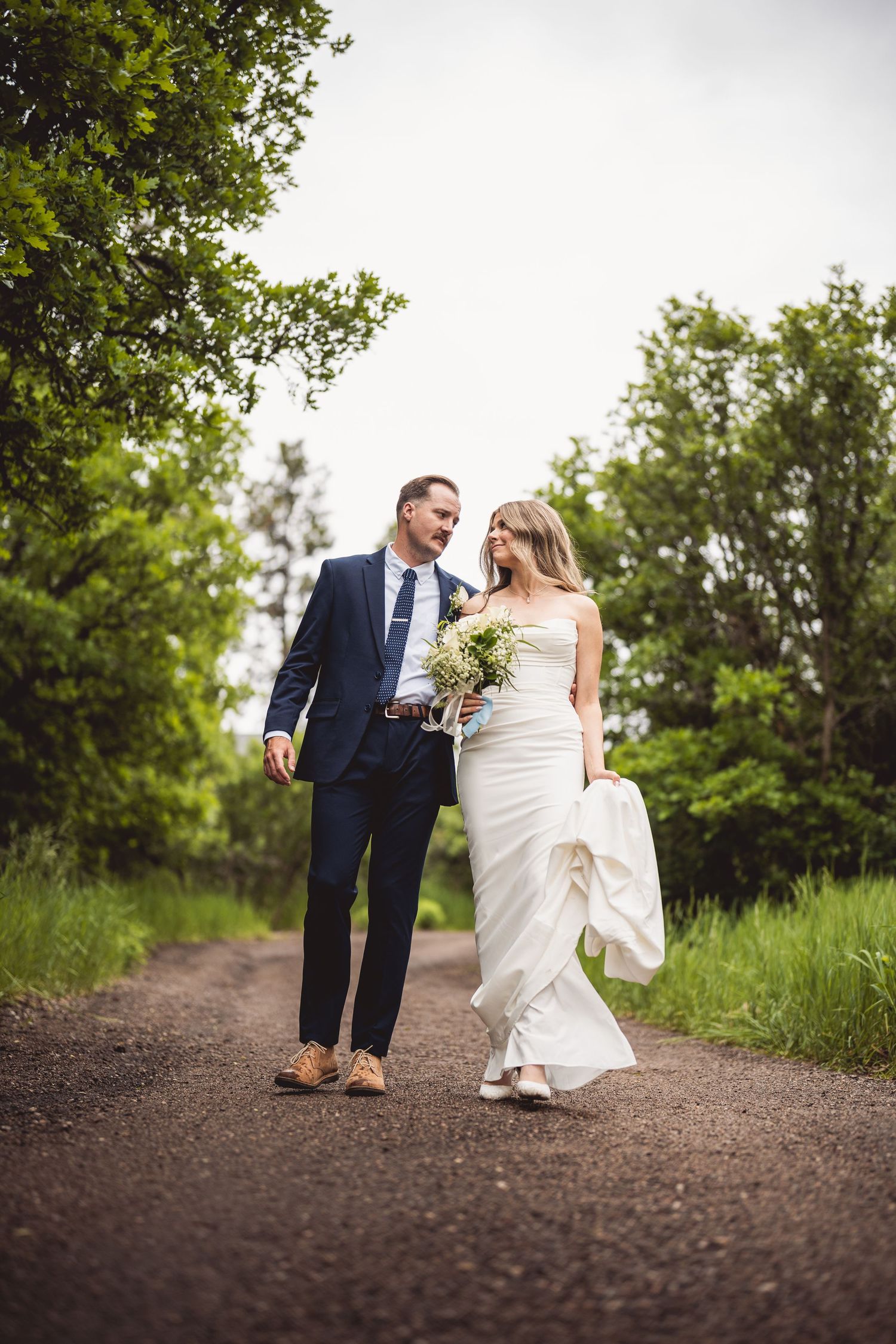 Wedding couple walking together on a dirt path surrounded by green foliage.