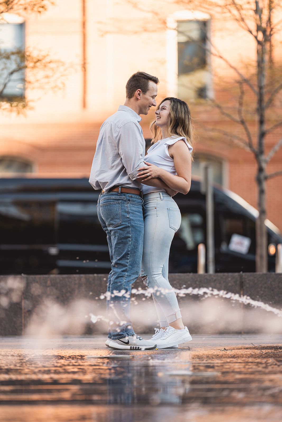 Couple shares intimate moment with splashing water at sunset against urban backdrop.