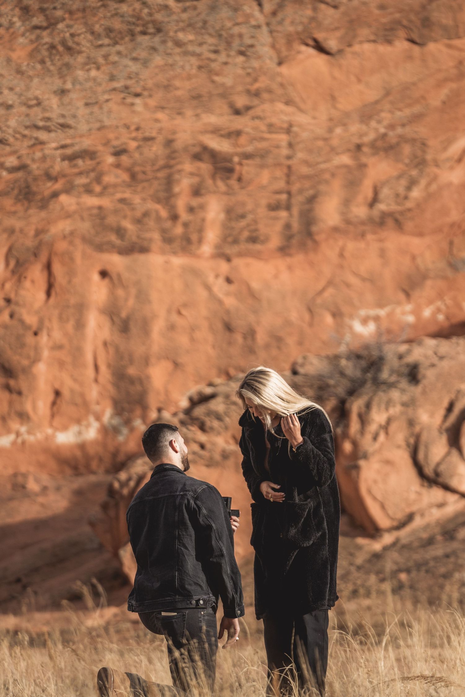 A couple walks together against striking red rock formations in a desert landscape.