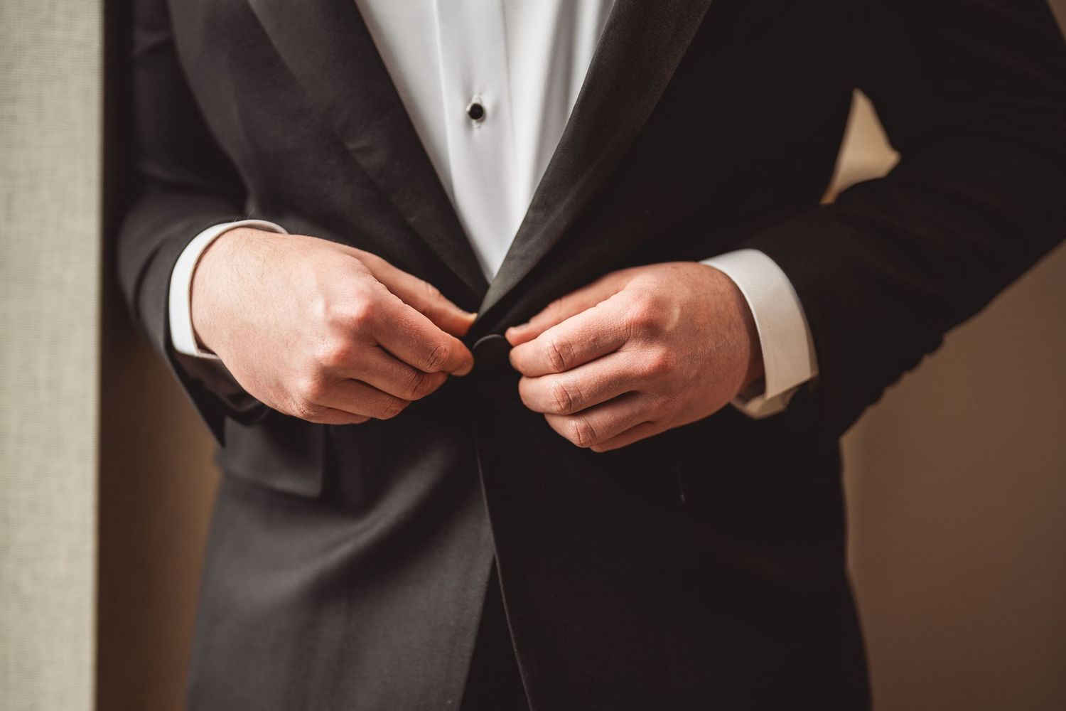 Close-up of hands buttoning a black tuxedo jacket during formal wedding preparation.